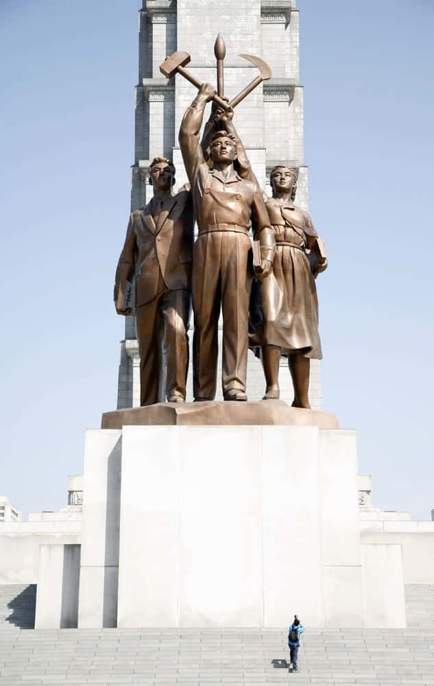 North Korea 桌布 壁紙 — 三個青銅工人手持鎚子與鐮刀，站在白色基座上，背景是清澈的藍天，基座底部有一個小人作為比例參考。