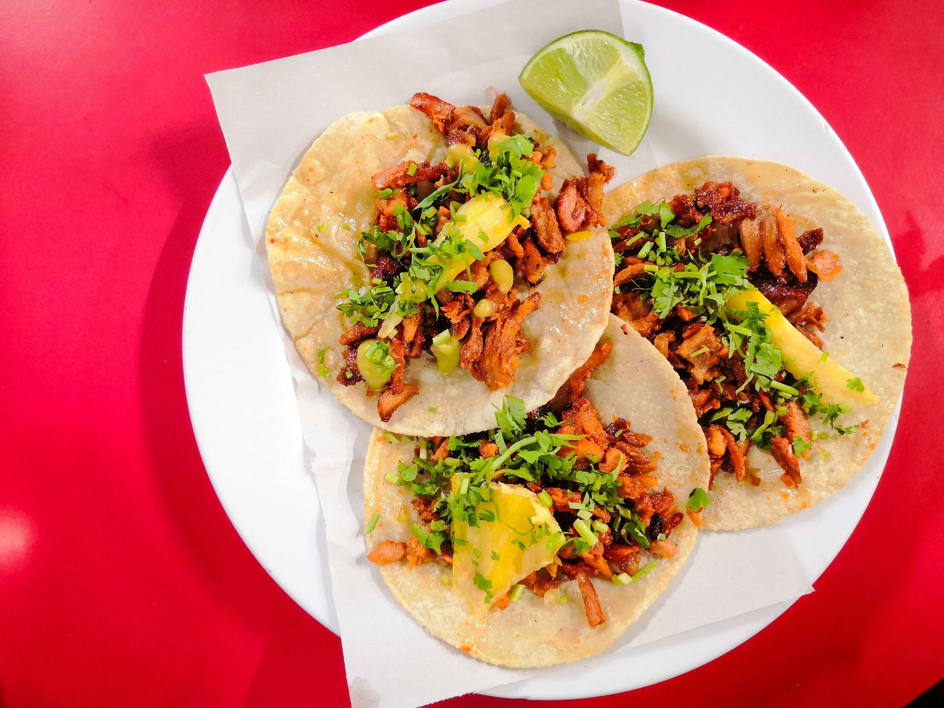 Mexican Food 桌布 壁紙 — 三個香脆墨西哥捲餅，上面鋪有撕碎的肉、酪梨和香菜，放在白色盤子上，襯托著大膽的紅色背景。