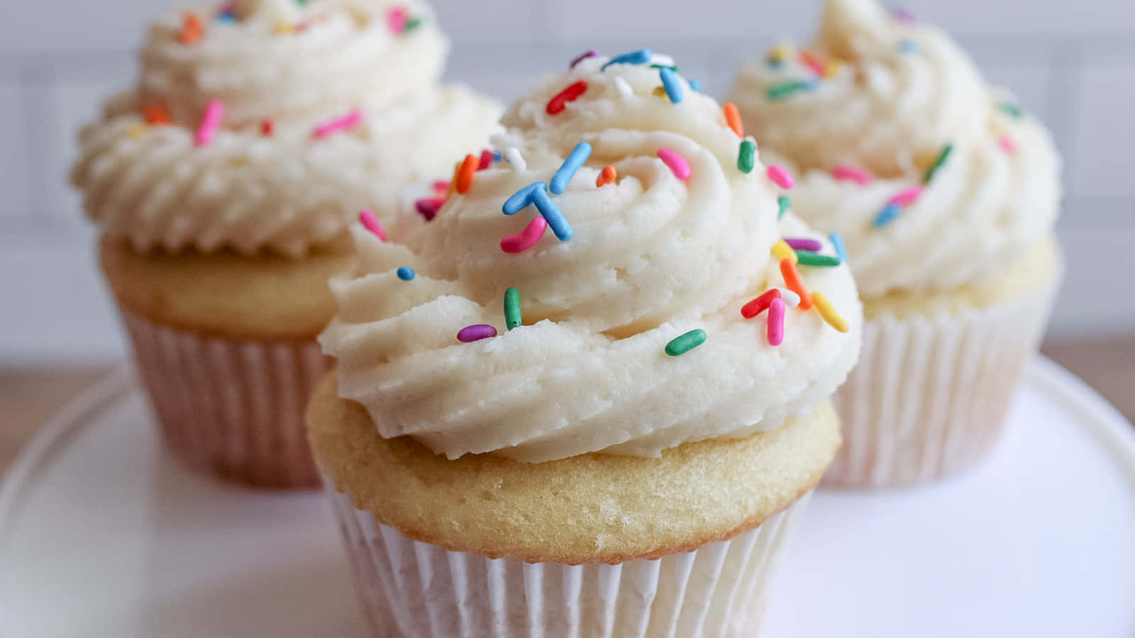 Buttercream 桌布 壁紙 — 三個香草紙杯蛋糕，上面有奶油糖霜和彩色糖粒，放在白色盤子上。