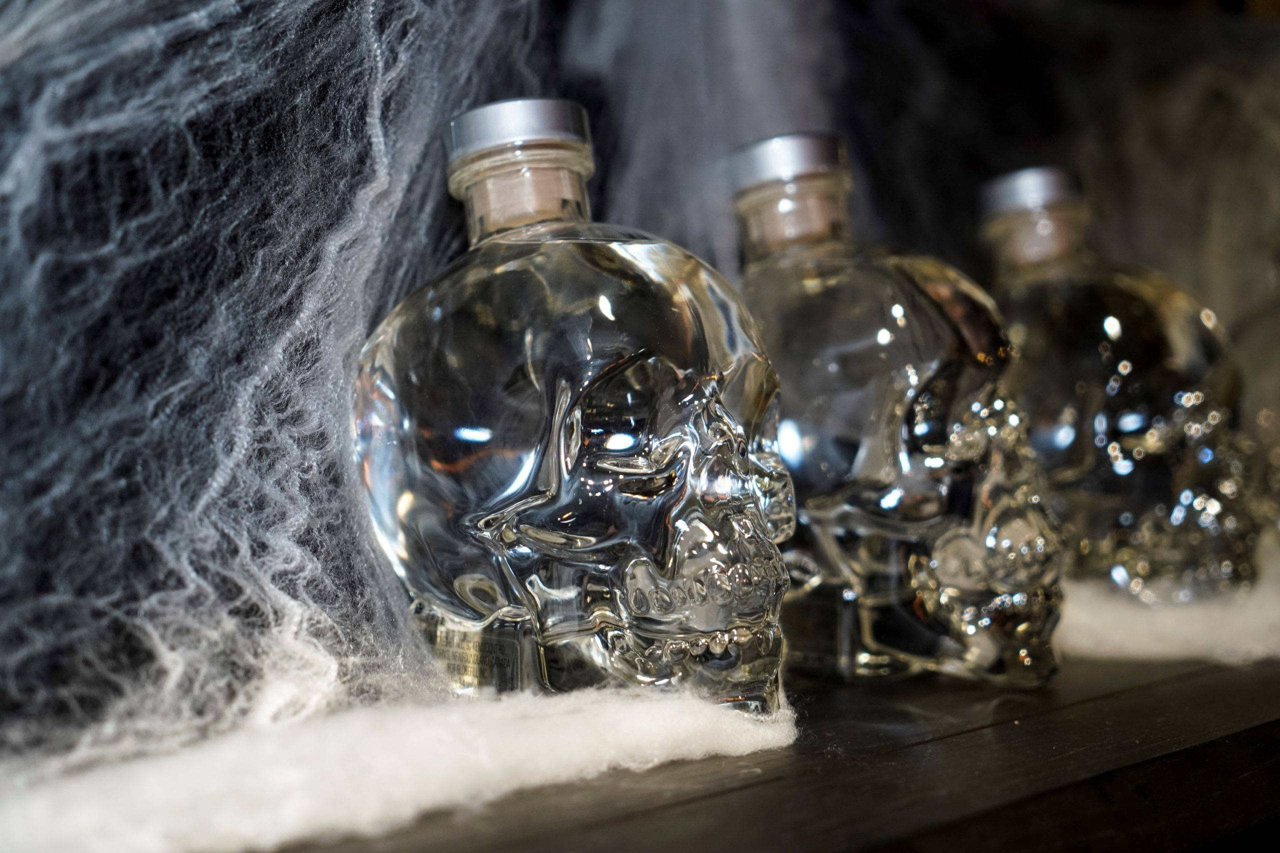 Drink 桌布 壁紙 — 三個骷髏形狀的玻璃伏特加酒瓶在黑暗的架子上閃閃發光，旁邊有縷縷白色蜘蛛網和霜。