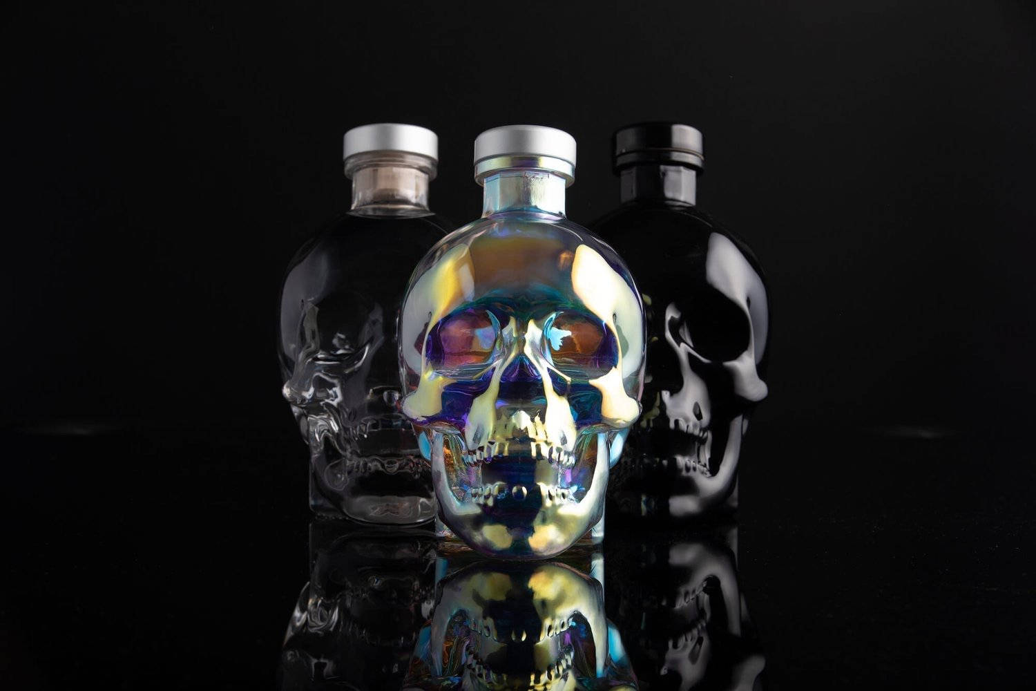 Drink 桌布 壁紙 — 三個骷髏形狀的酒瓶，帶有彩虹色和啞光飾面，立在反射黑色表面上，背景為深色。