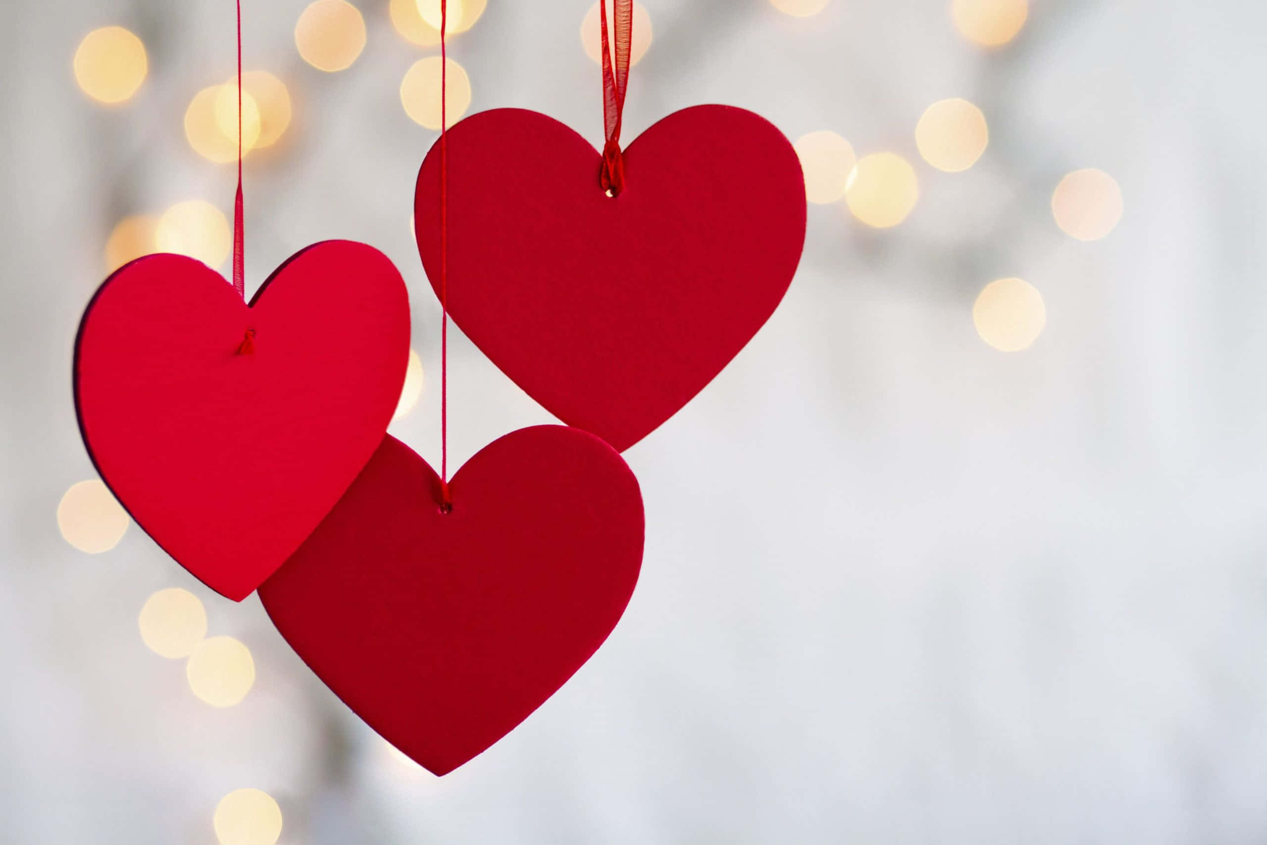 Happy Valentines Day 桌布 壁紙 — 三個鮮紅色的紙愛心懸掛在柔和的白色背景上，伴隨著溫暖的金色散景燈光。