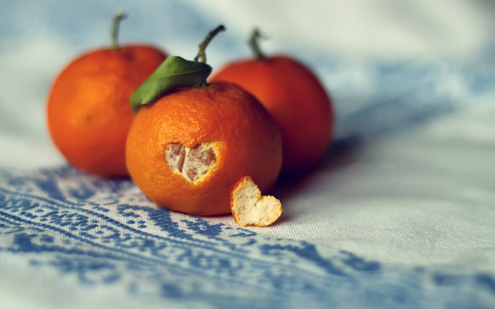 Fruits 桌布 壁紙 — 三個鮮豔的柳橙放在藍色圖案布上，其中一個被咬成心形，露出多汁的果肉。