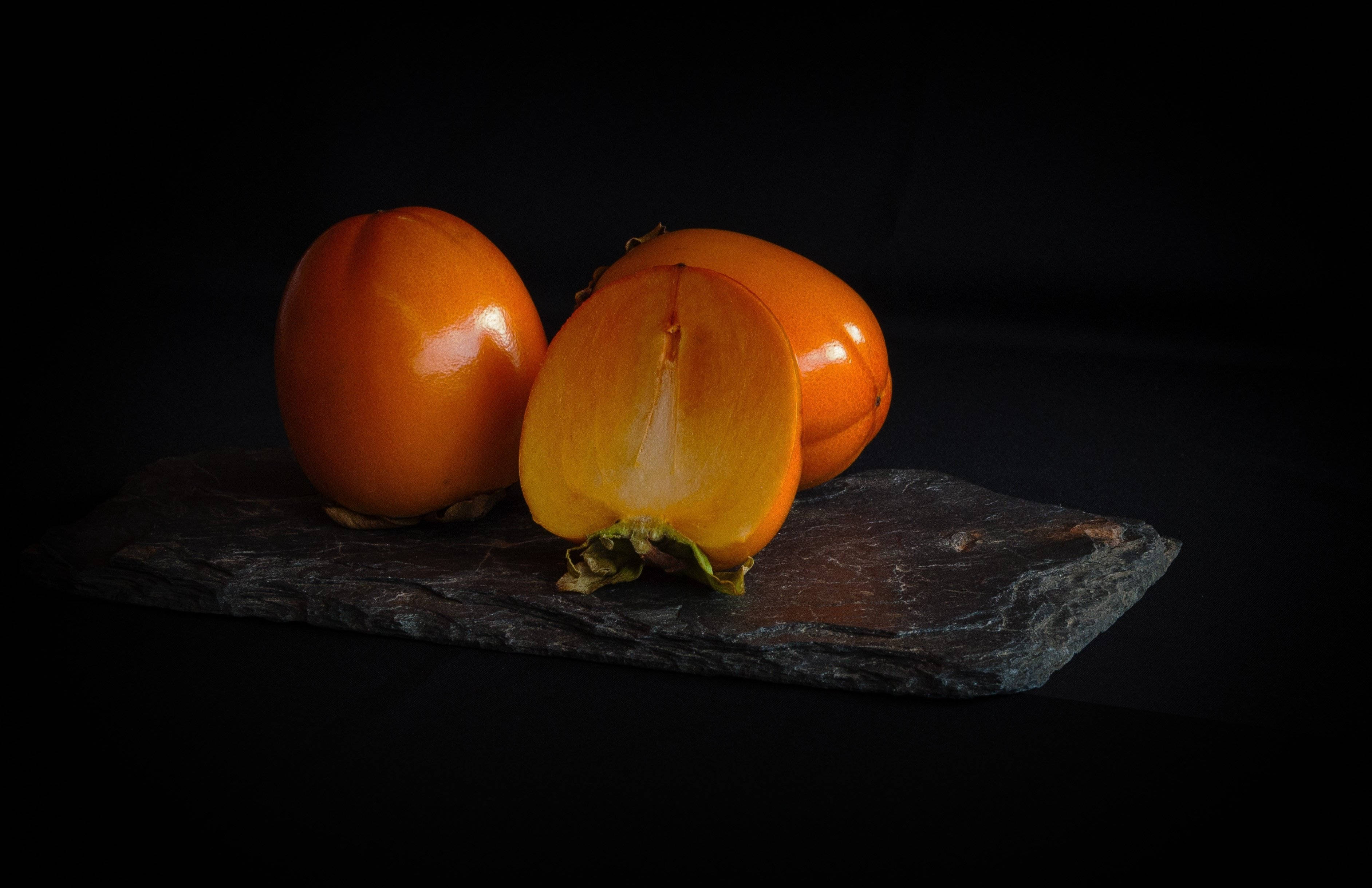 Fruit 桌布 壁紙 — 三個鮮豔的橙色柿子，其中一個切成兩半，放在黑色背景的深色石板板上。