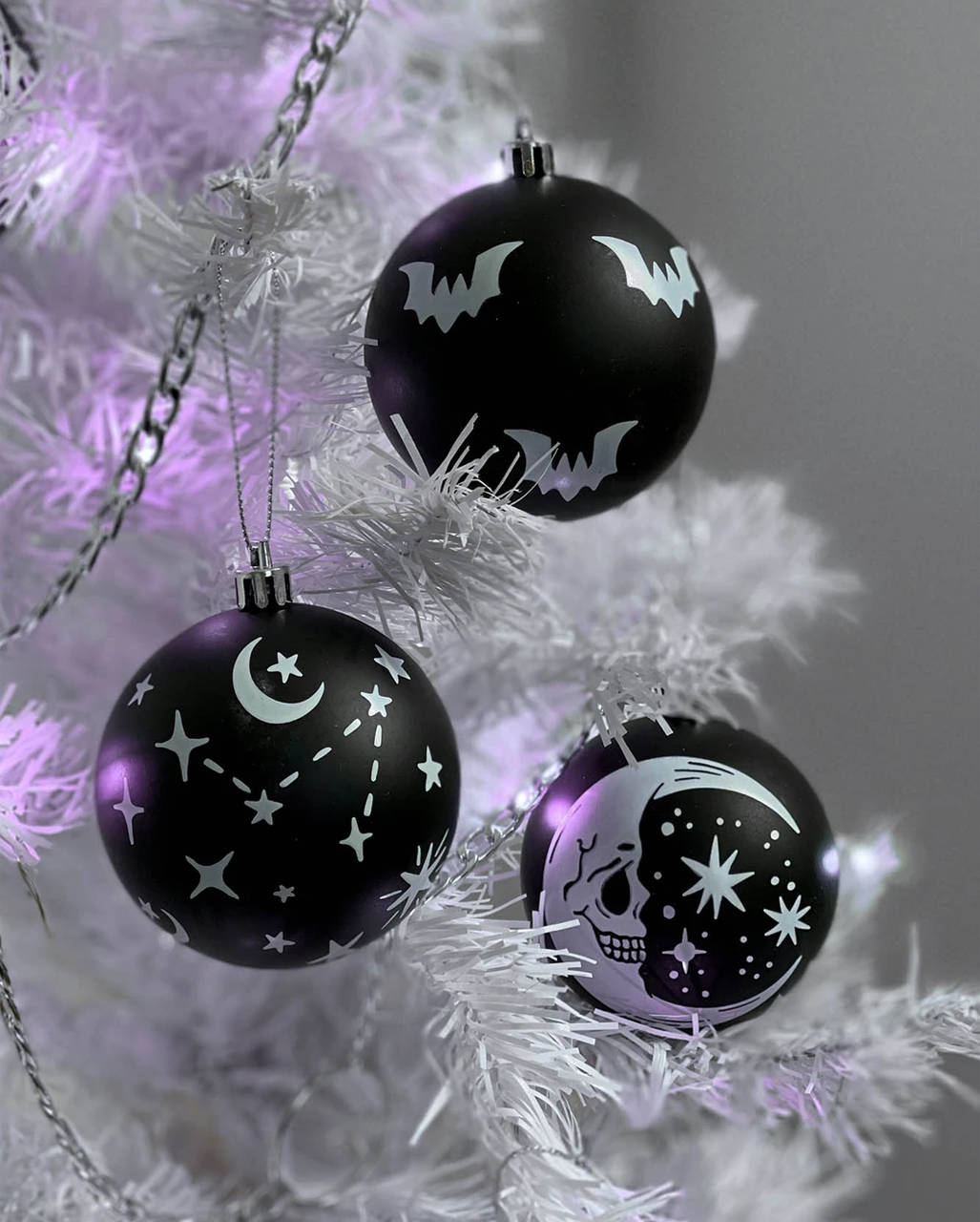 Gothic Christmas 桌布 壁紙 — 三個黑色萬聖節裝飾品，上面有蝙蝠、月亮、骷髏頭和星星，掛在帶有紫色燈光的白色樹上。