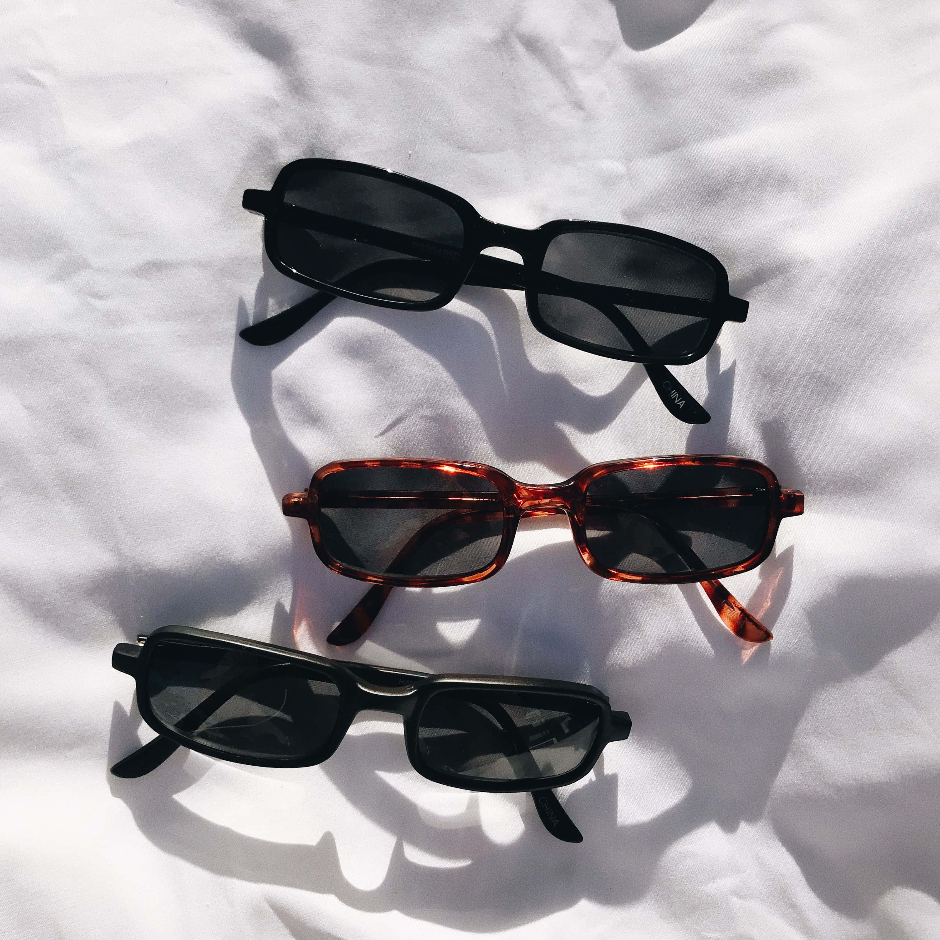 Sunglasses 桌布 壁紙 — 三副黑色玳瑁和黑色的長方形太陽眼鏡在明亮的陽光下放在皺巴巴的白色布料上。