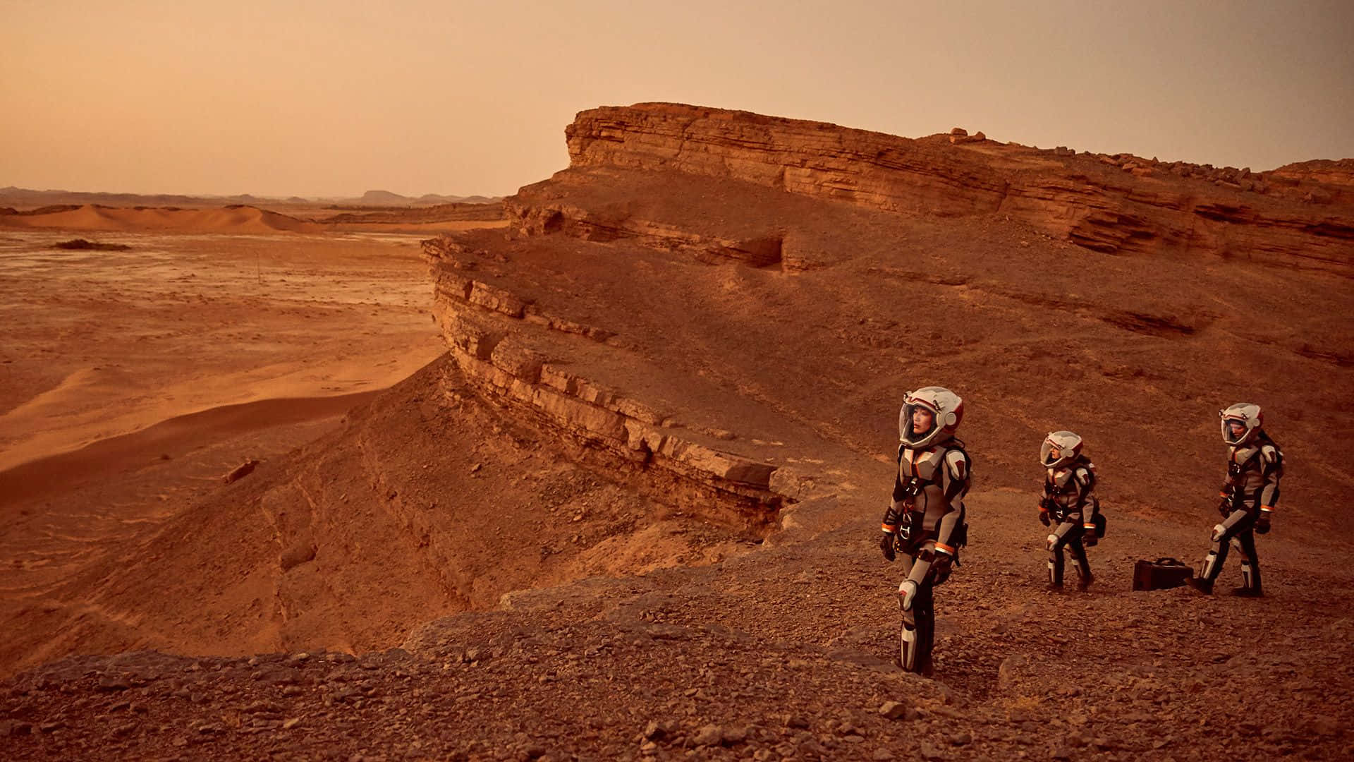 Mars Landscape 桌布 壁紙 — 三名全副武裝的太空人在朦朧的橙色天空下探索崎嶇的紅色火星峽谷。