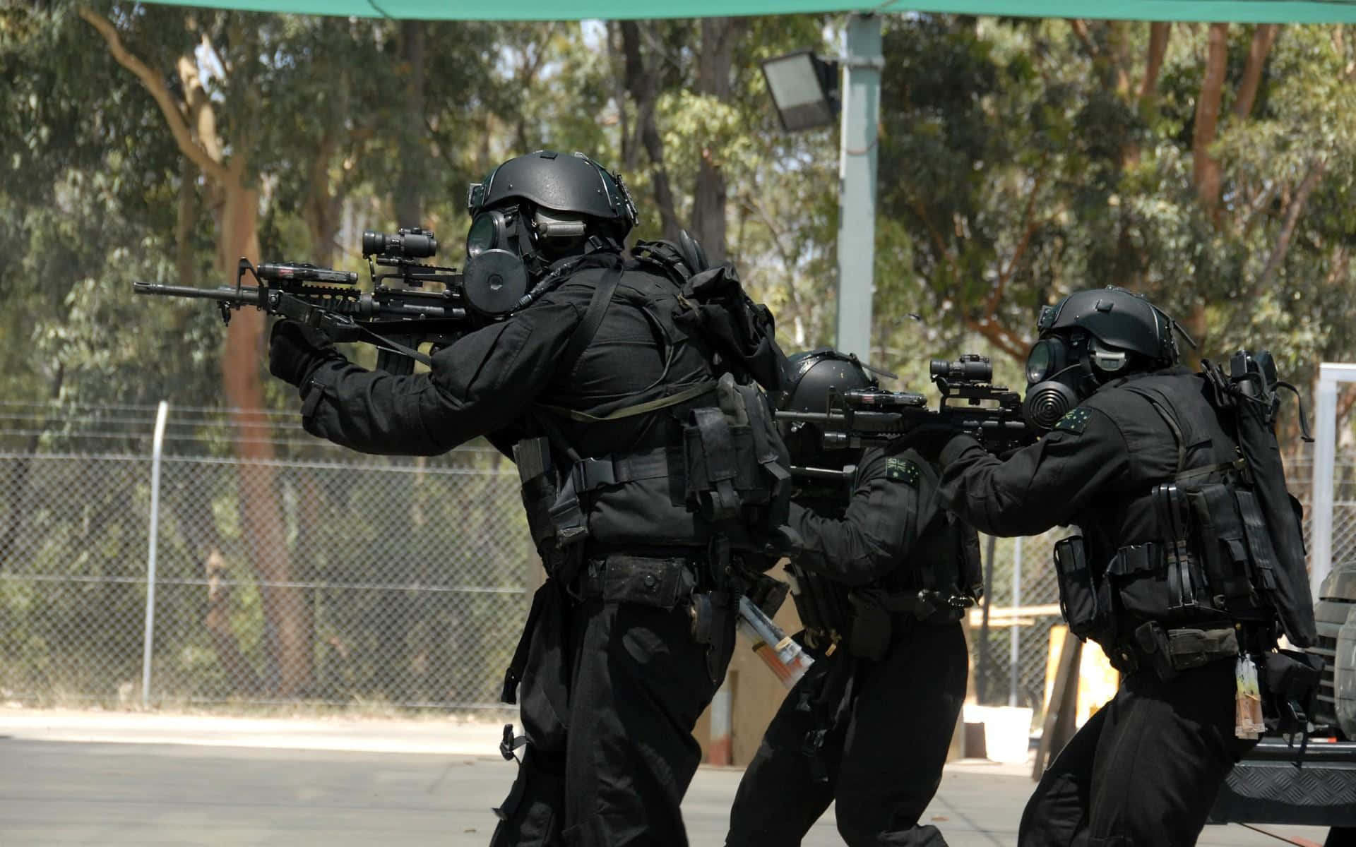Swat 桌布 壁紙 — 三名全副武裝的戰術人員持步槍穿過陽光普照的戶外訓練區，周圍有樹木和圍欄。