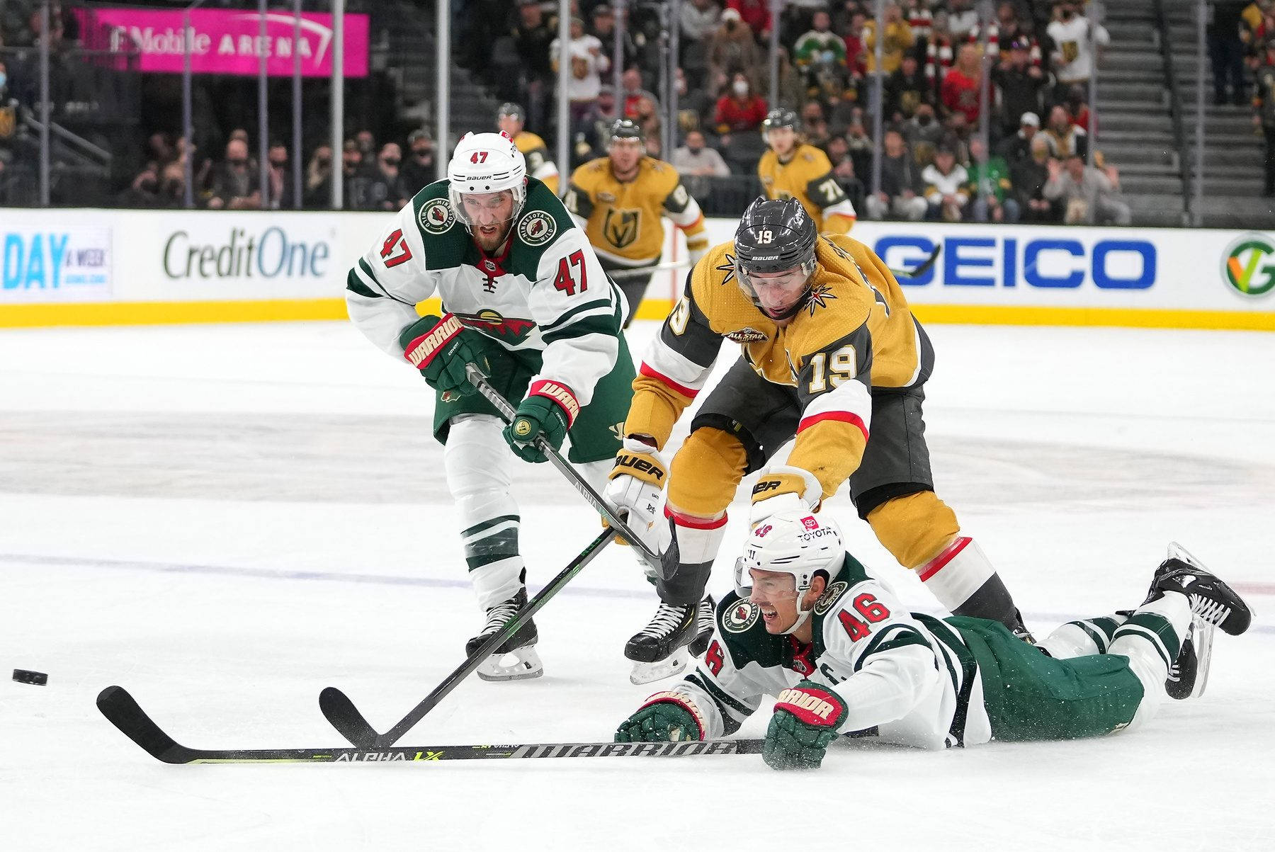 hockey 桌布 壁紙 — 三名冰球運動員在冰上激烈爭奪，其中一人趴著滑行，觀眾在看台上觀看。
