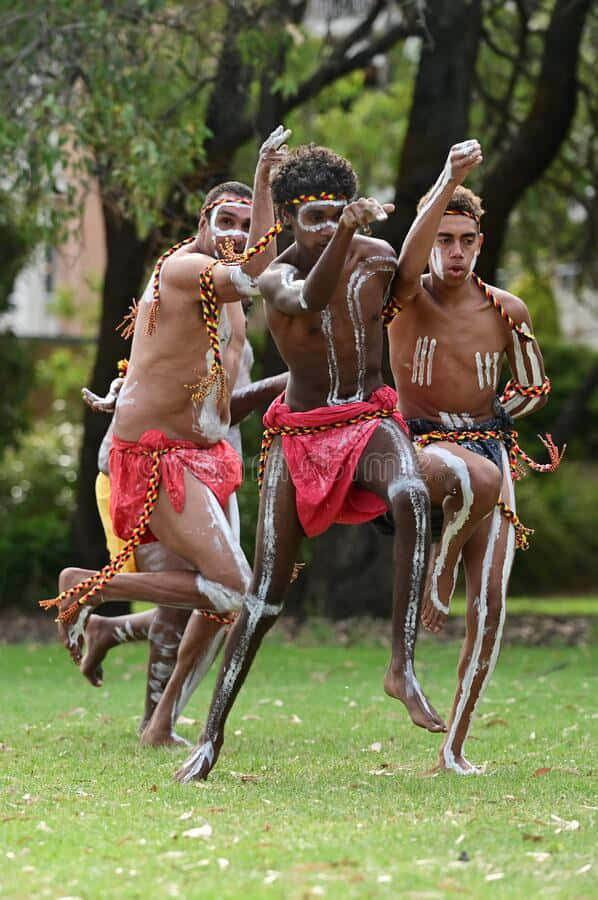 Aboriginal 桌布 壁紙 — 三名原住民男子身穿傳統身體彩繪和紅色腰布，在明亮的日光下動態地跳過草地公園。