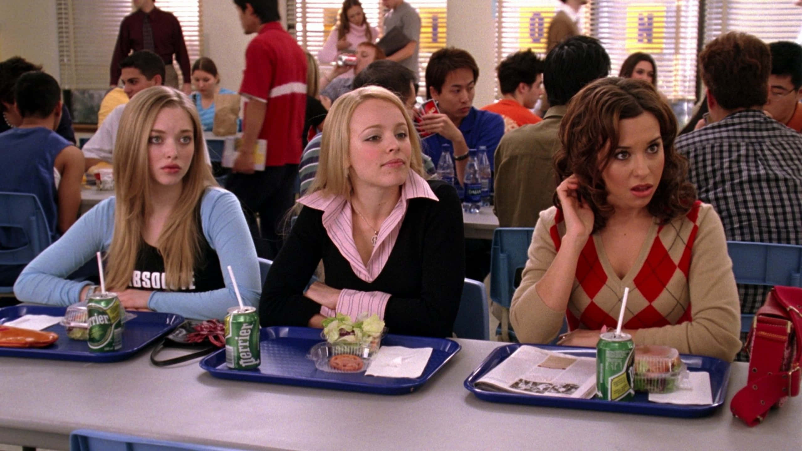 mean girls 桌布 壁紙 — 三名少女坐在學校食堂的餐桌旁，面前放著裝有食物和飲料的托盤，表情驚訝或擔憂。