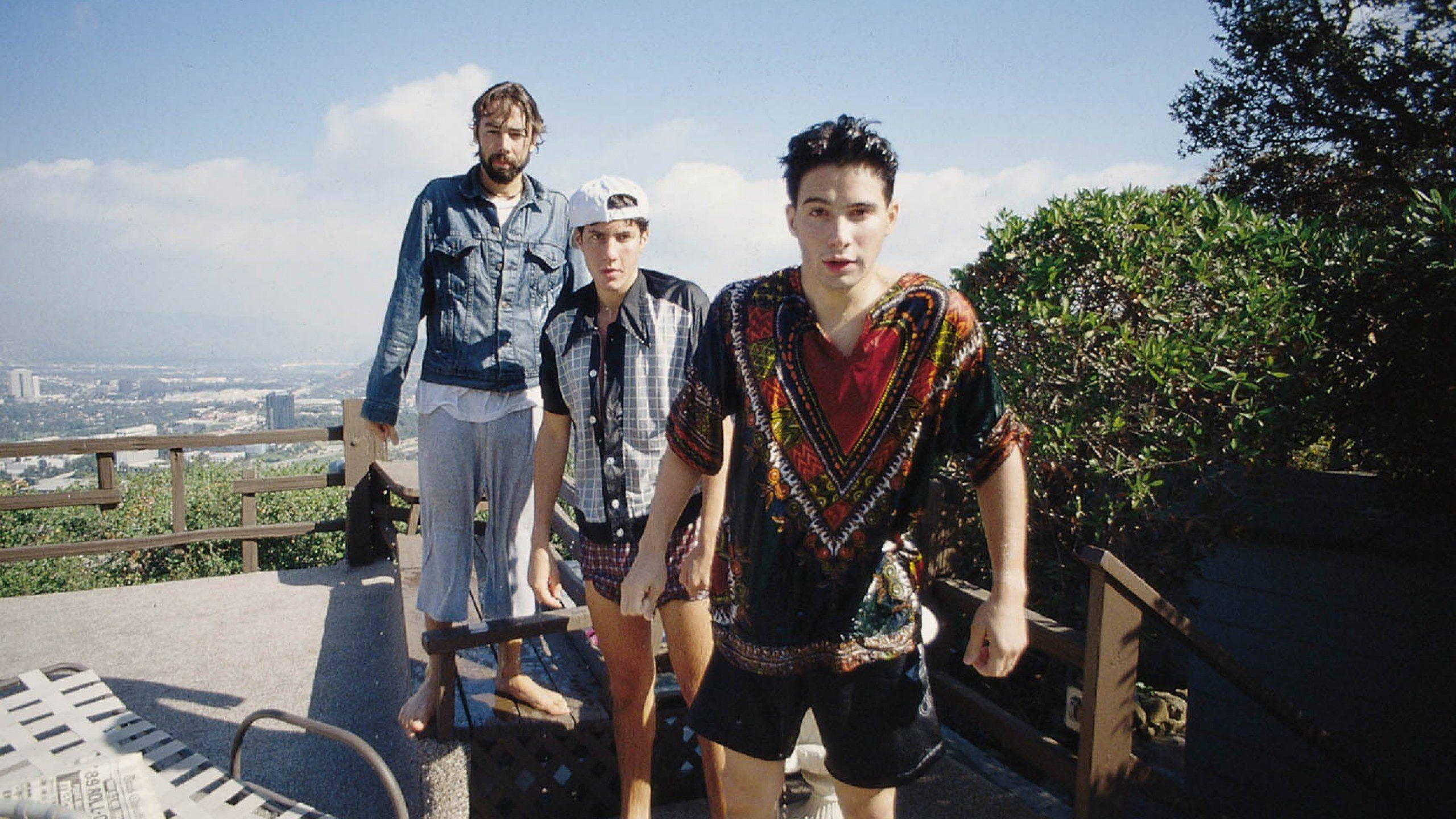 Rappers 桌布 壁紙 — 三名年輕男子穿著休閒夏季服裝，站在山坡上，俯瞰著晴朗藍天下的一座廣闊城市。