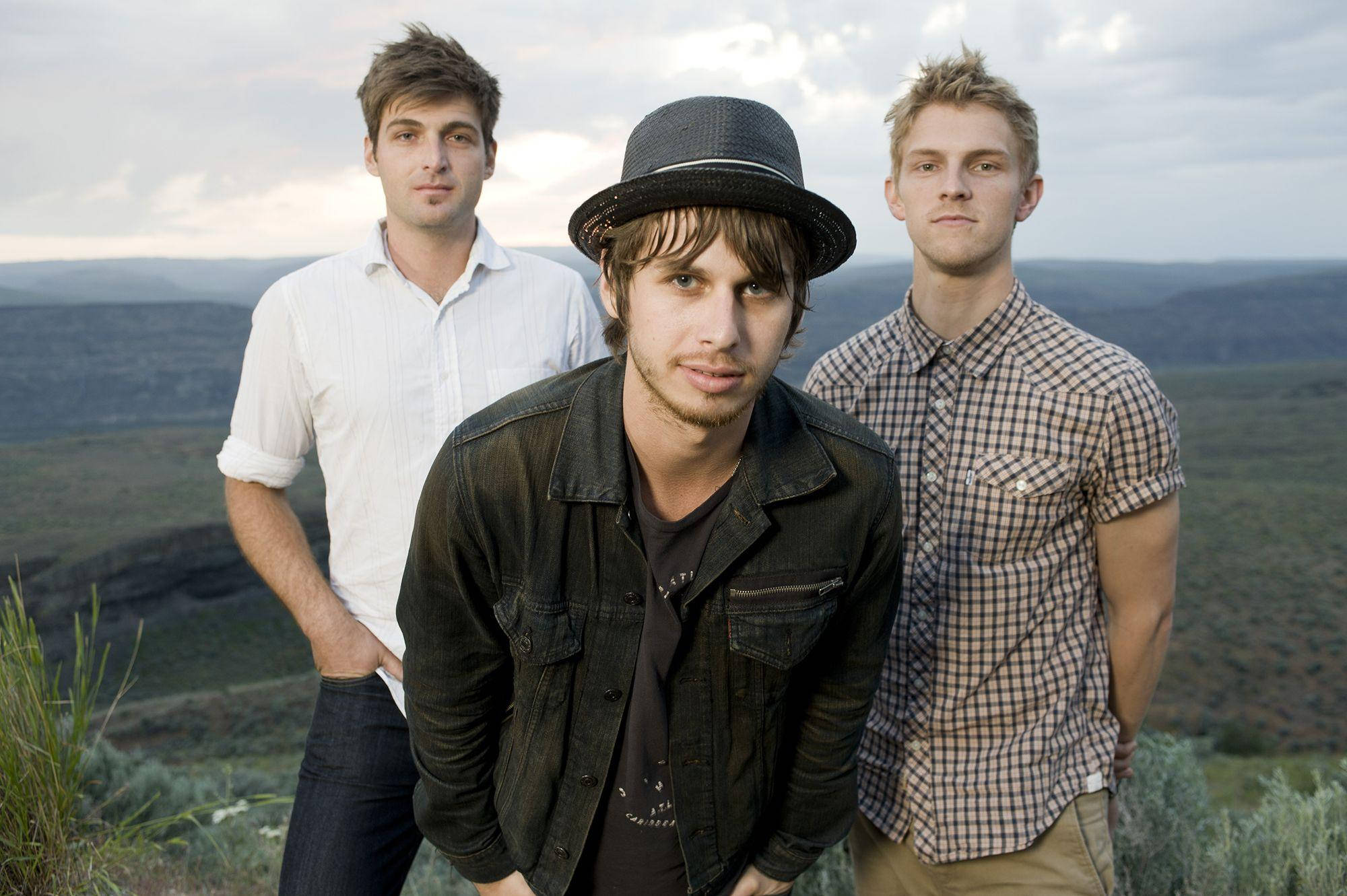 foster the people 桌布 壁紙 — 三名年輕男子自信地在長滿青草的山坡上擺姿勢，俯瞰著多雲天空下廣闊的丘陵景觀。