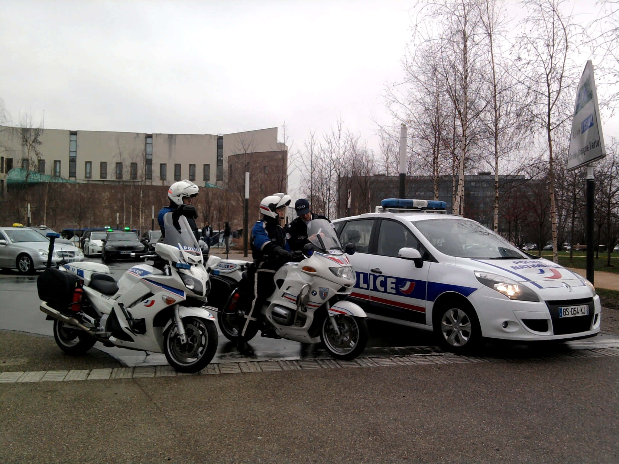Law Enforcement 桌布 壁紙 — 三名戴白色頭盔的警察站在兩輛摩托車和一輛有標記的警車旁邊，地點是濕漉漉的城市人行道，附近有光禿禿的樹木。