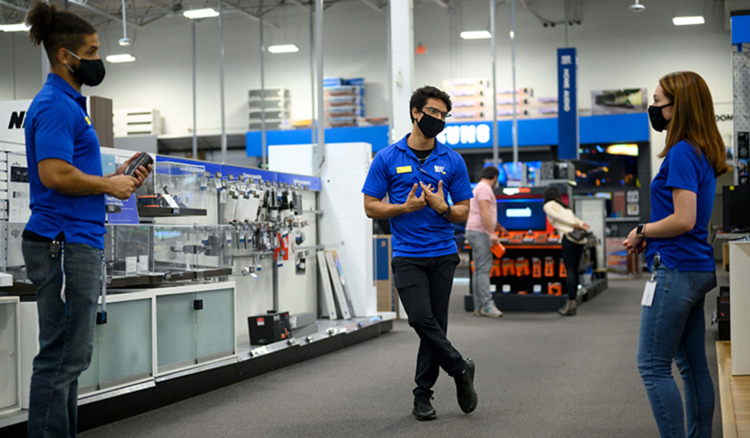 Best Buy 桌布 壁紙 — 三名戴著口罩、身穿藍色襯衫的員工在光線充足、擺滿工具和用品的五金店通道上交談。