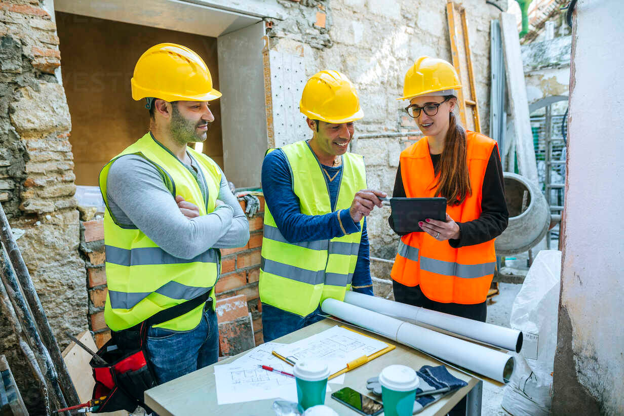 construction 桌布 壁紙 — 三名戴著黃色安全帽和反光背心的建築工人在一個雜亂的工地桌子上，周圍是磚塊和工具，正在審閱藍圖。