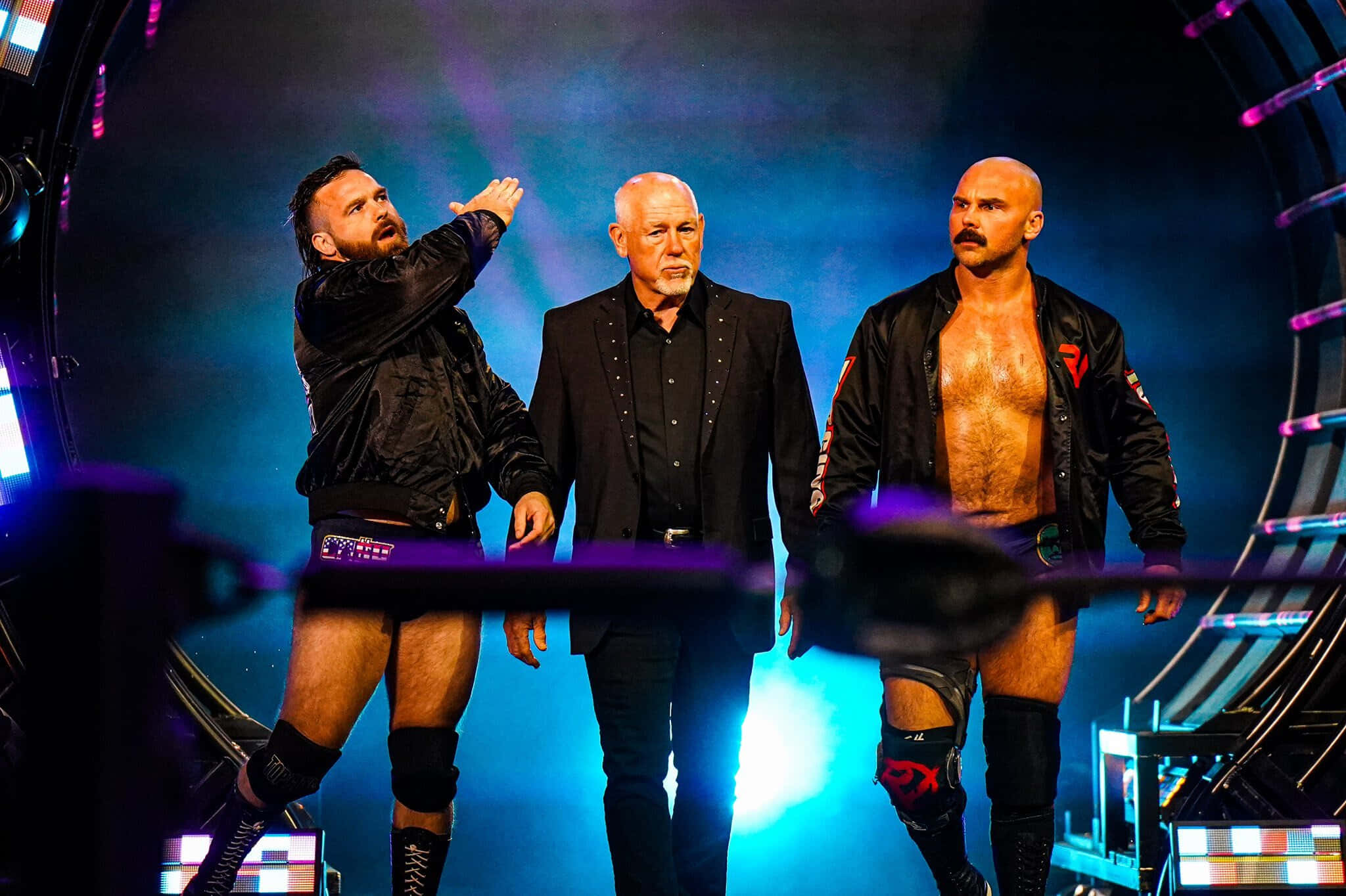 WWE Desktop 桌布 壁紙 — 三名摔角手和一名穿西裝的男子在充滿活力的藍色和紫色舞台燈光下戲劇性地站立。