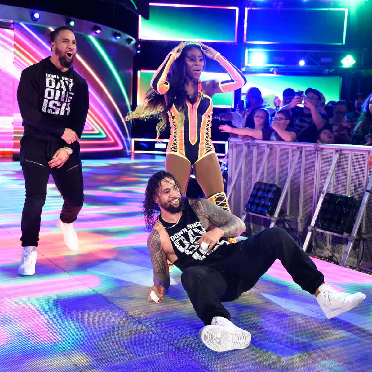 WWE 桌布 壁紙 — 三名摔角手在充滿活力的霓虹燈舞台上精力充沛地表演，背後有歡呼的人群隔著屏障。