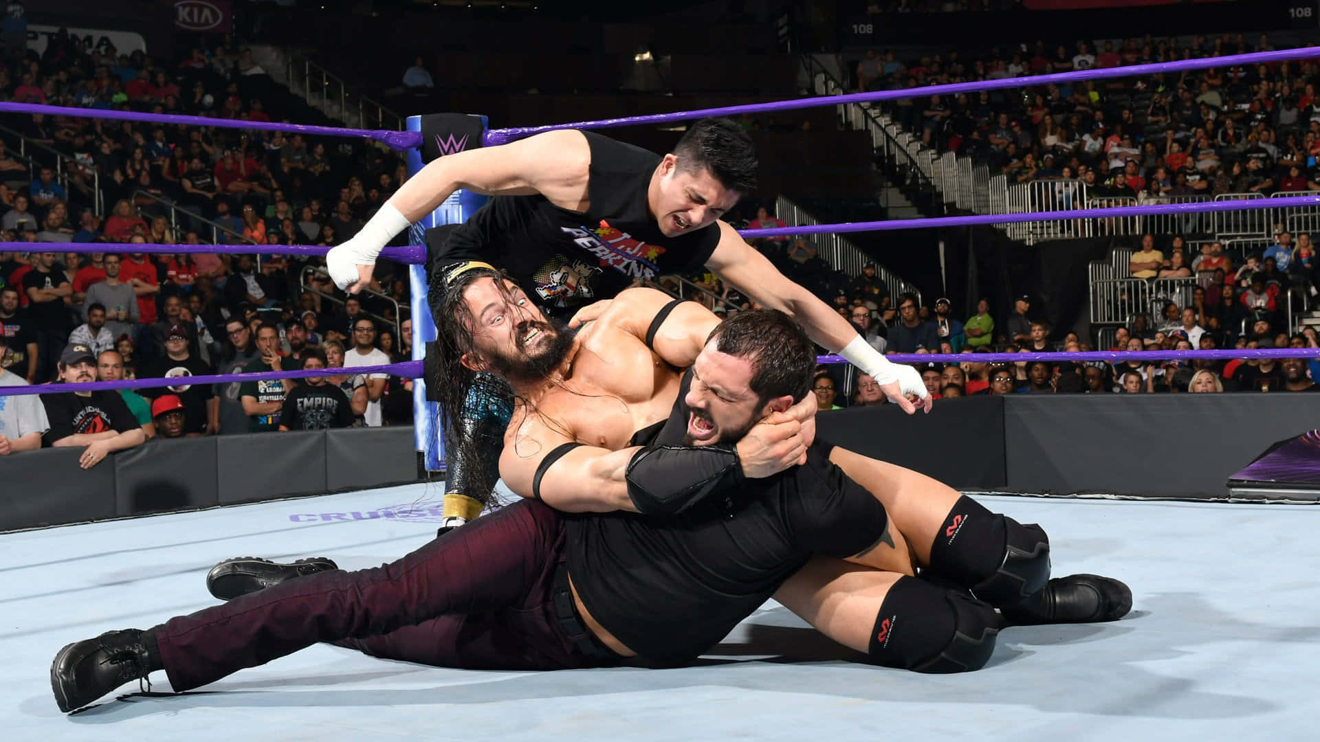 WWE 桌布 壁紙 — 三名摔角手在擂臺上激烈纏鬥，全場觀眾在明亮的場館燈光下觀看。