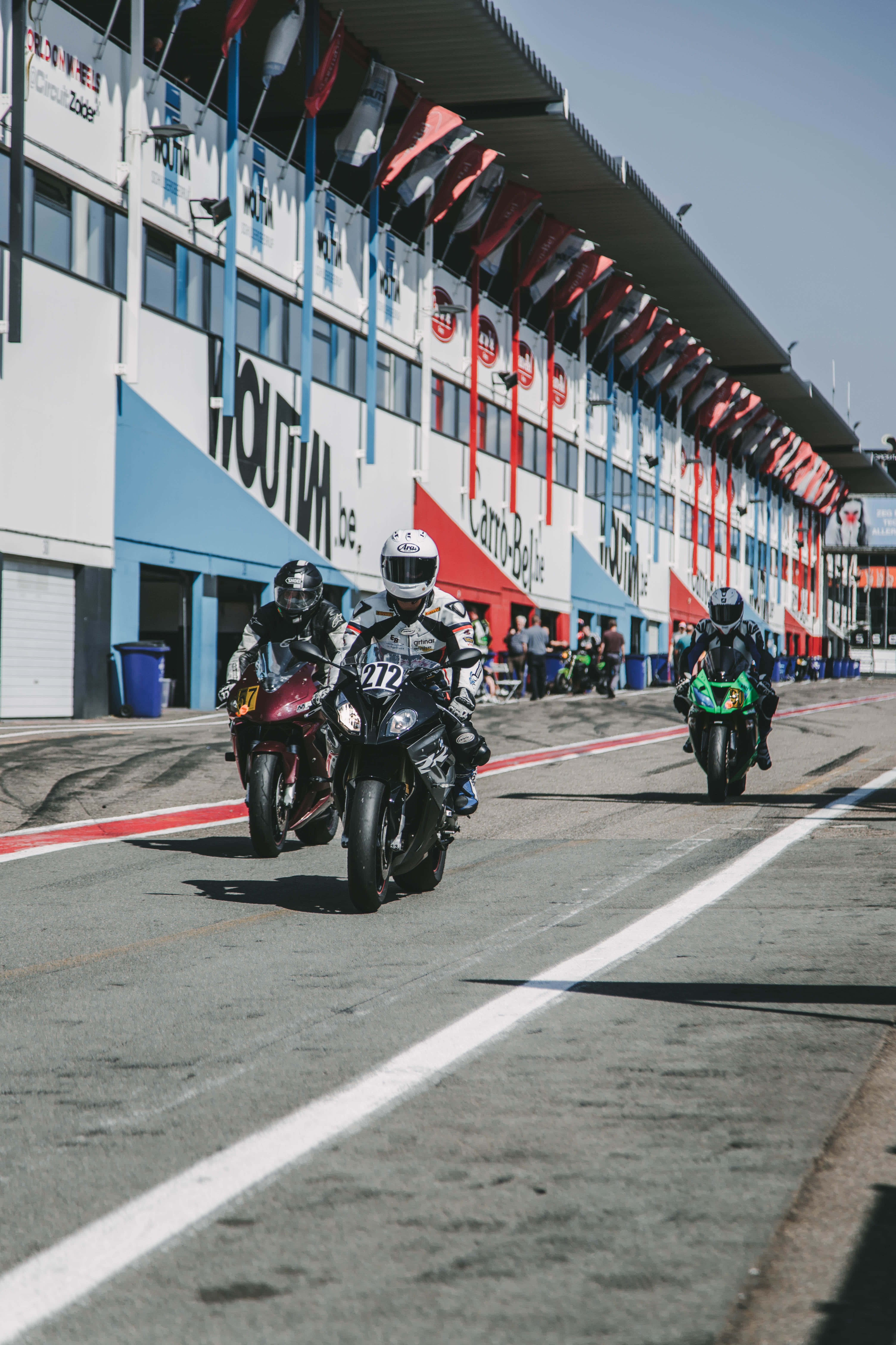 Race Track 桌布 壁紙 — 三名摩托車手在陽光明媚的賽道上比賽，經過一個色彩繽紛的維修站建築物，上面掛有贊助商橫幅。