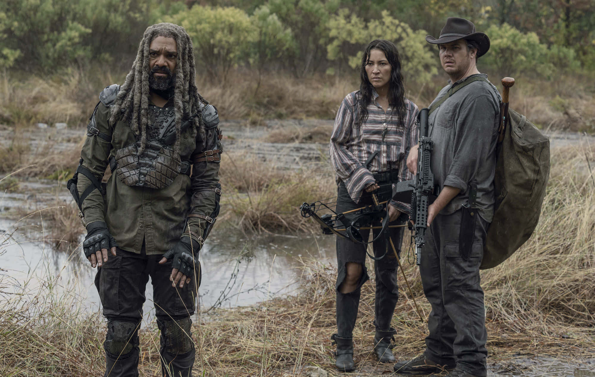 walking dead 桌布 壁紙 — 三名武裝倖存者在一片泥濘的田野中保持警覺，身後有茂密的植被和淺溪。