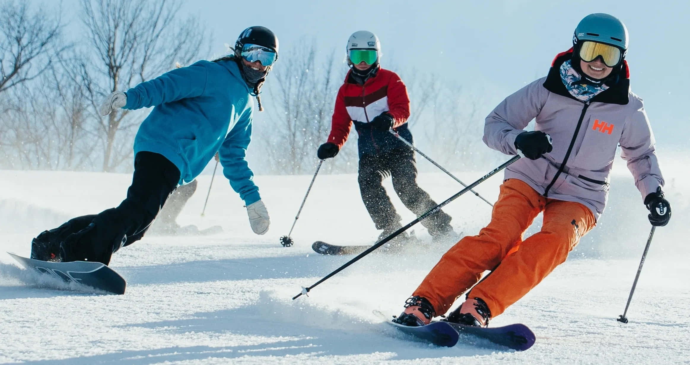 ski 桌布 壁紙 — 三名滑雪者在雪坡上刻劃轉彎，在晴朗的藍天下揚起粉雪，背景是光禿禿的樹木。