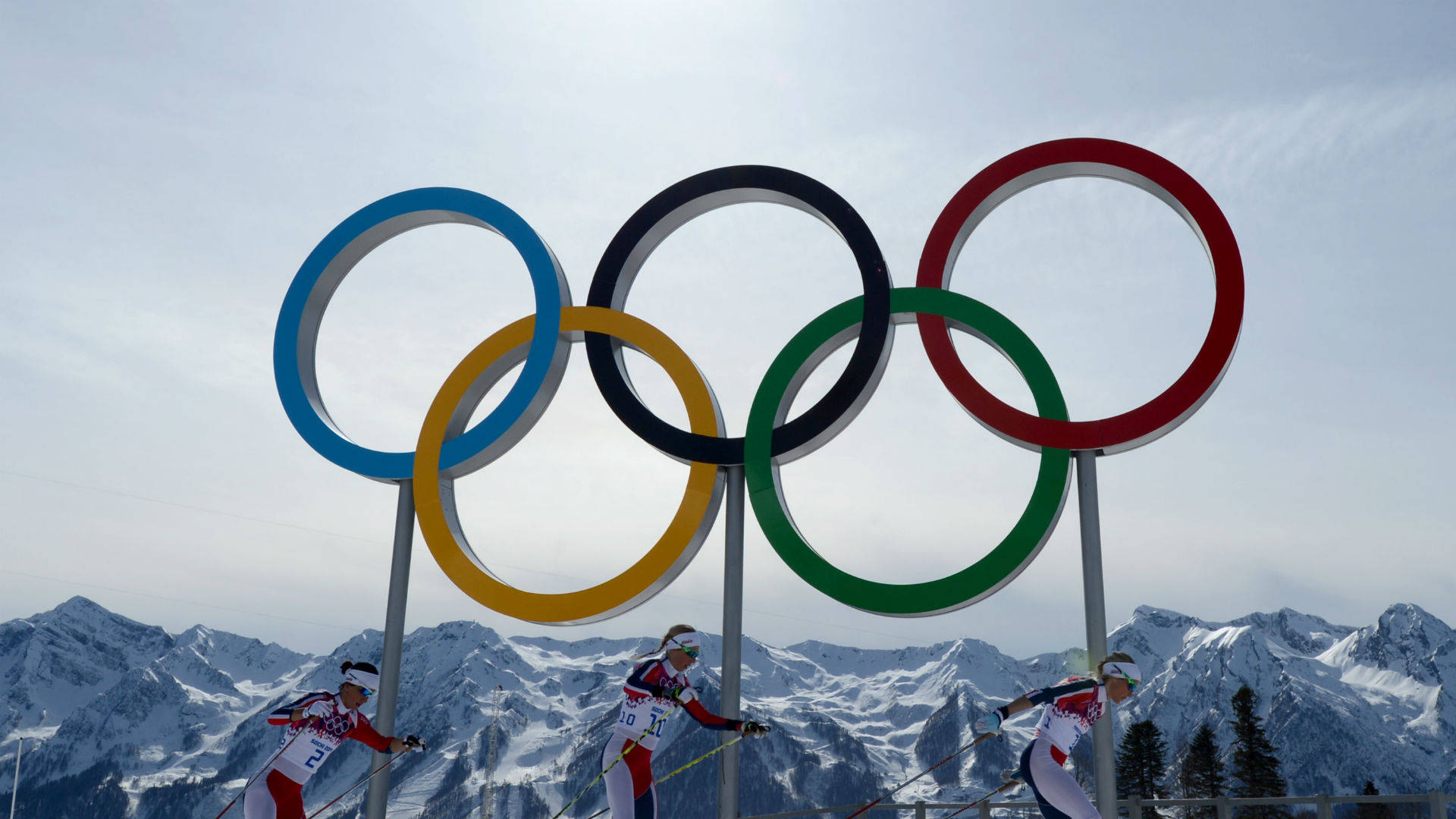 Winter Olympics 桌布 壁紙 — 三名滑雪選手在蒼白的天空下，以積雪覆蓋的山脈為背景，飛馳而過巨大的奧運五環。