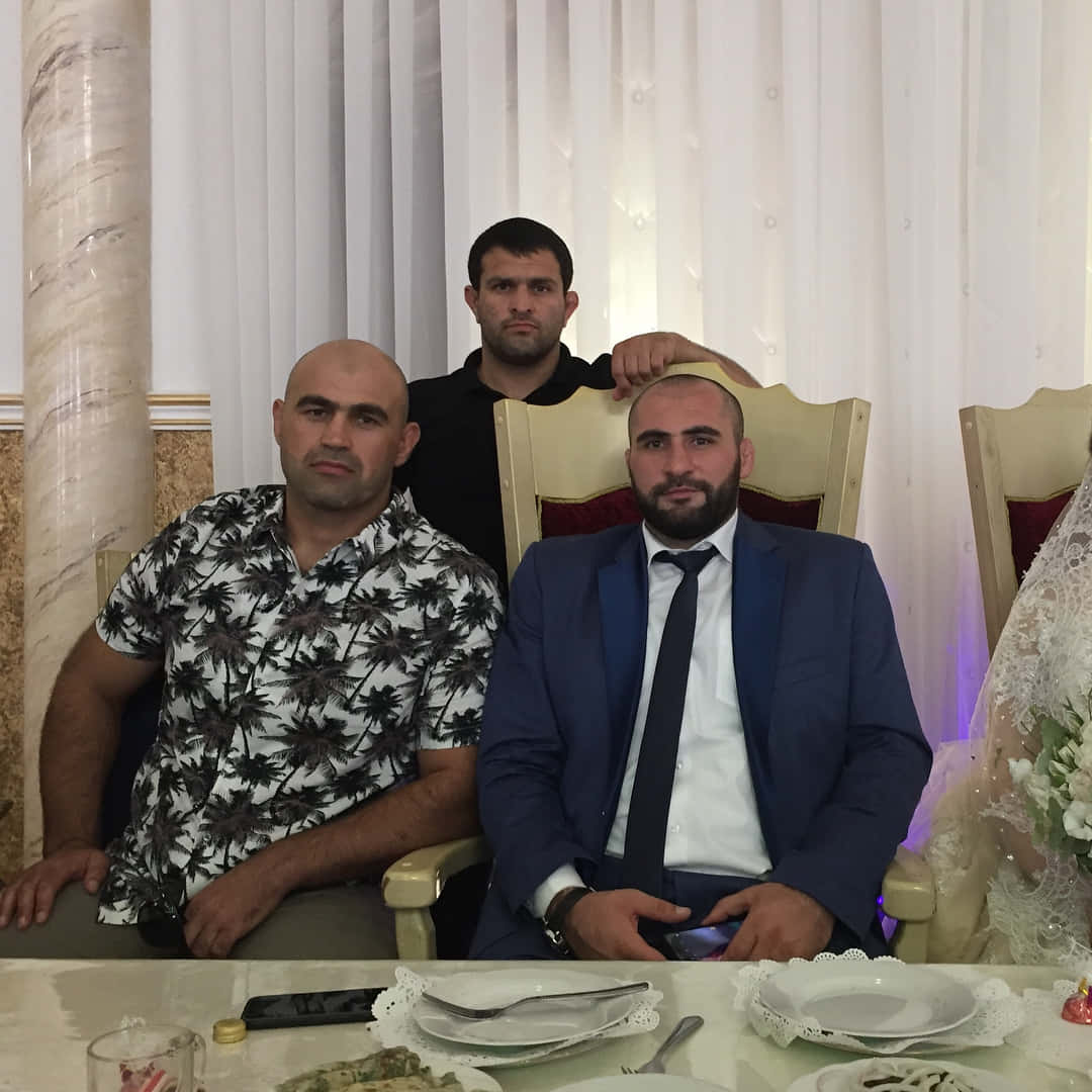 Wedding 桌布 壁紙 — 三名男士在裝飾華麗的宴會桌前正式擺姿勢，背景有白色蕾絲盤子和大理石柱。