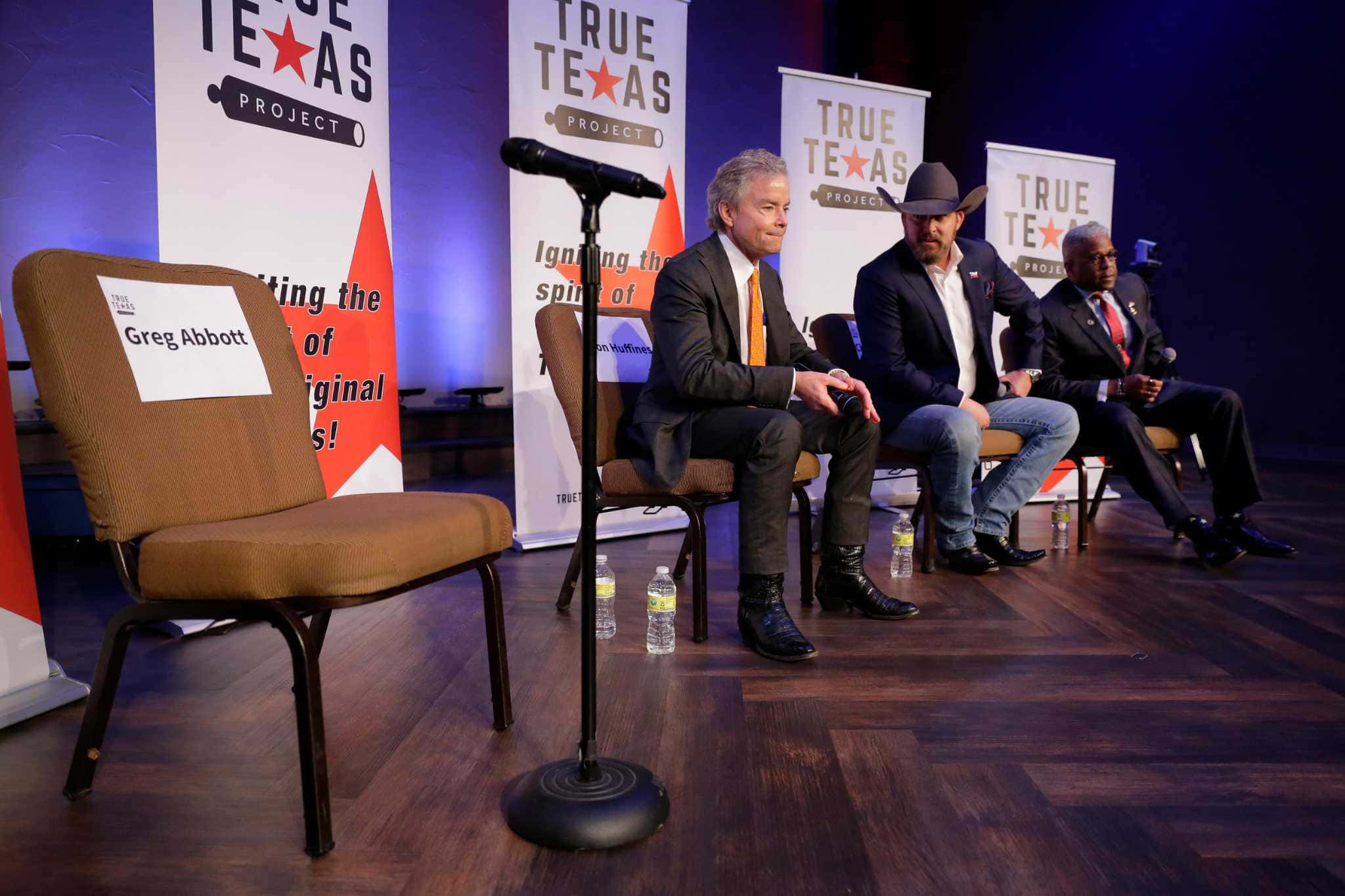 Media 桌布 壁紙 — 三名男子在「True Texas Project」活動的舞台上就座，前景有一張標有「Greg Abbott」的空椅子。