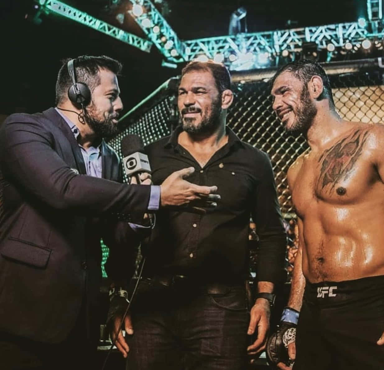 MMA 桌布 壁紙 — 三名男子在UFC八角籠中，一名採訪者手持麥克風和耳機，兩名拳手，一名赤裸上身帶刺青微笑