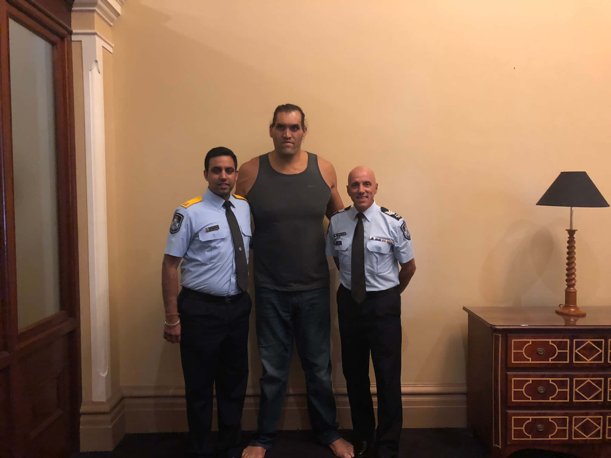 Indian 桌布 壁紙 — 三名男子在室內站立，兩名穿制服的員警站在一名穿背心的高大男子兩側，背景是米色牆壁。