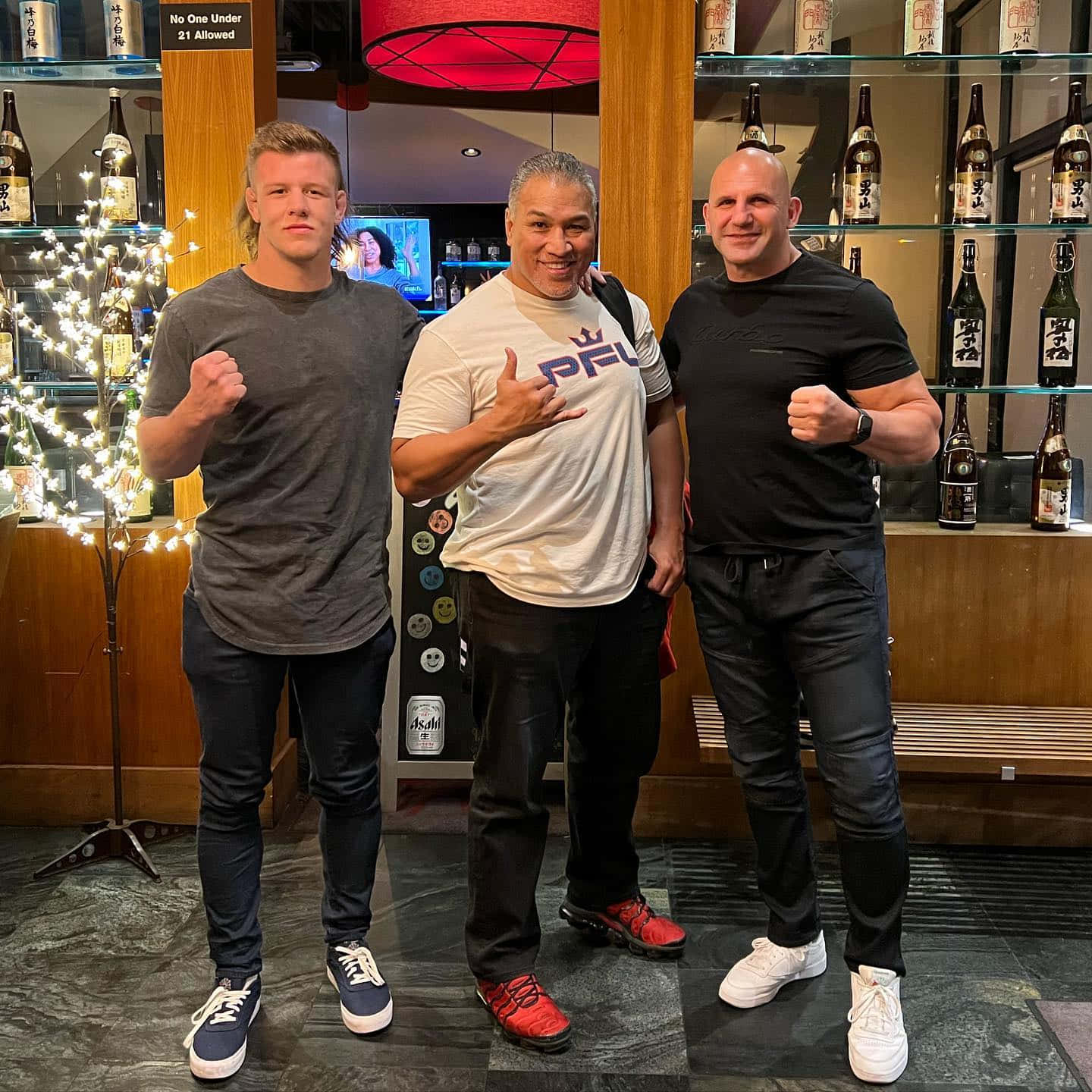 UFC 桌布 壁紙 — 三名男子在燈光昏暗的酒吧中自信地擺姿勢，身後有清酒瓶架和一棵發光的樹。