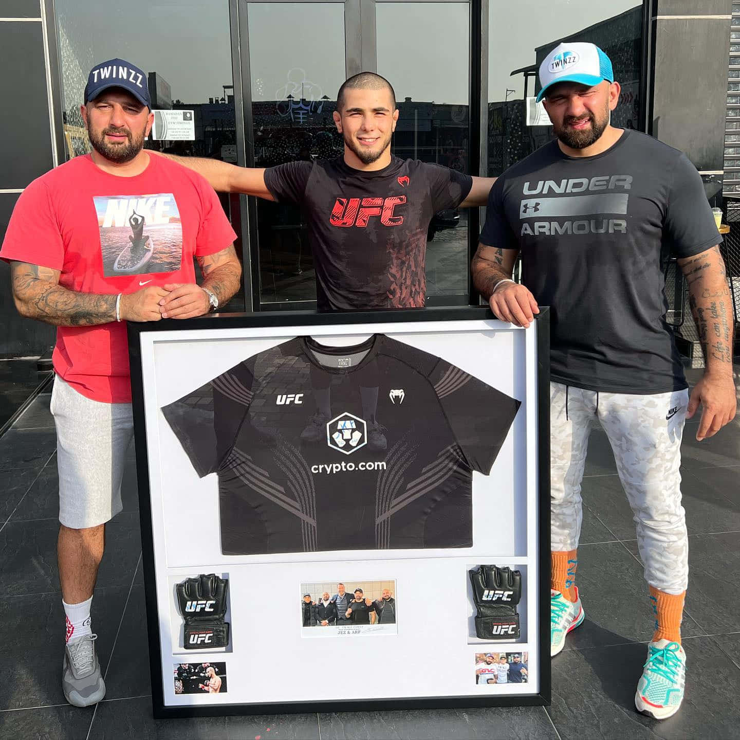 MMA 桌布 壁紙 — 三名男子在現代建築的玻璃入口旁，自豪地站在一個裱框的UFC拳手球衣和紀念品旁邊。