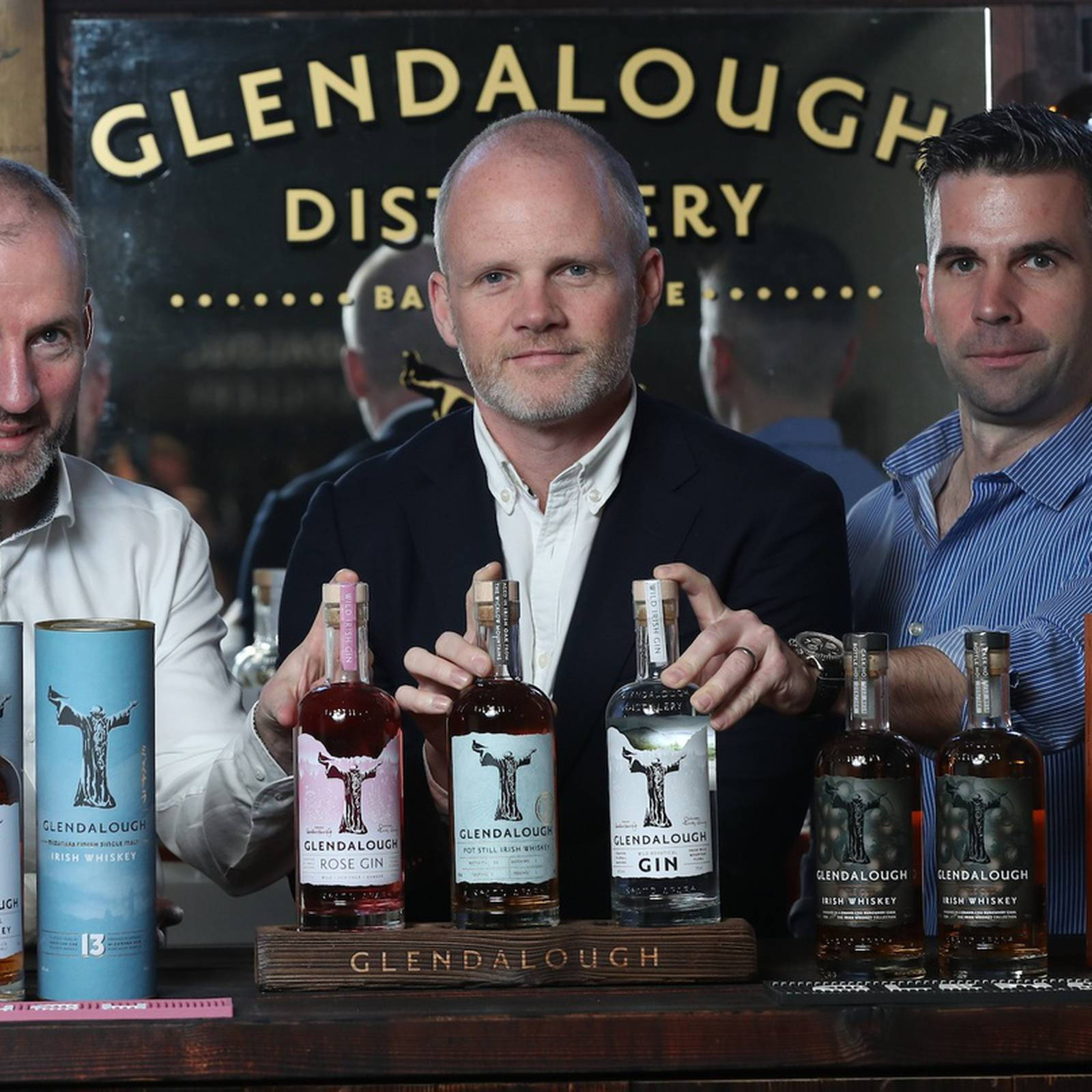 Whiskey 桌布 壁紙 — 三名男子在酒吧自豪地展示Glendalough Distillery烈酒，背景有品牌標誌和木製托盤。