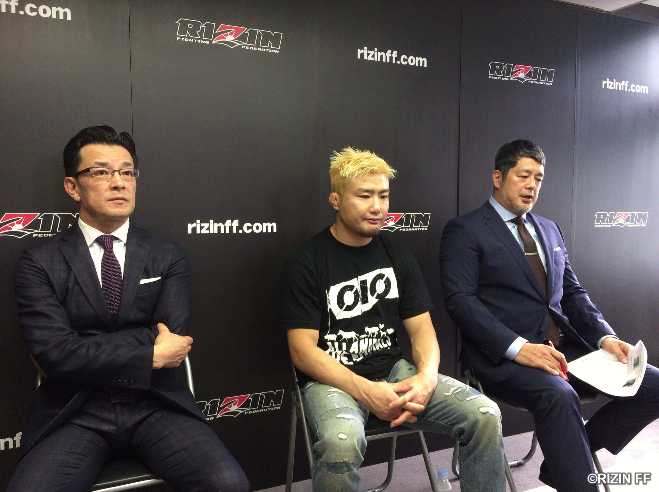 UFC 桌布 壁紙 — 三名男子坐在黑色Rizin FF品牌牆前，一人穿西裝，另一人穿休閒T恤和牛仔褲。