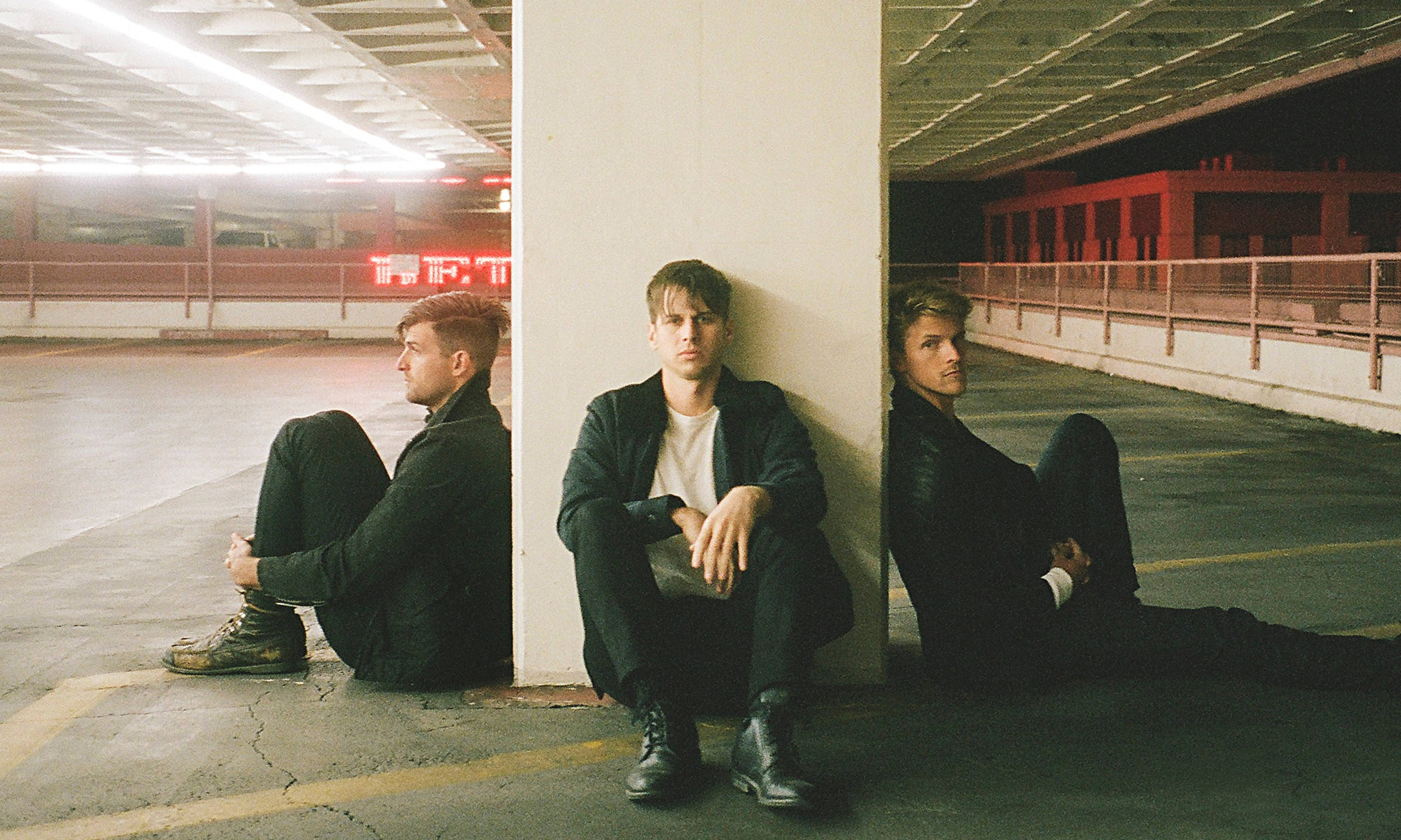 foster the people 桌布 壁紙 — 三名男子靠著柱子坐在光線昏暗的停車場，背景有紅色霓虹燈招牌閃爍。