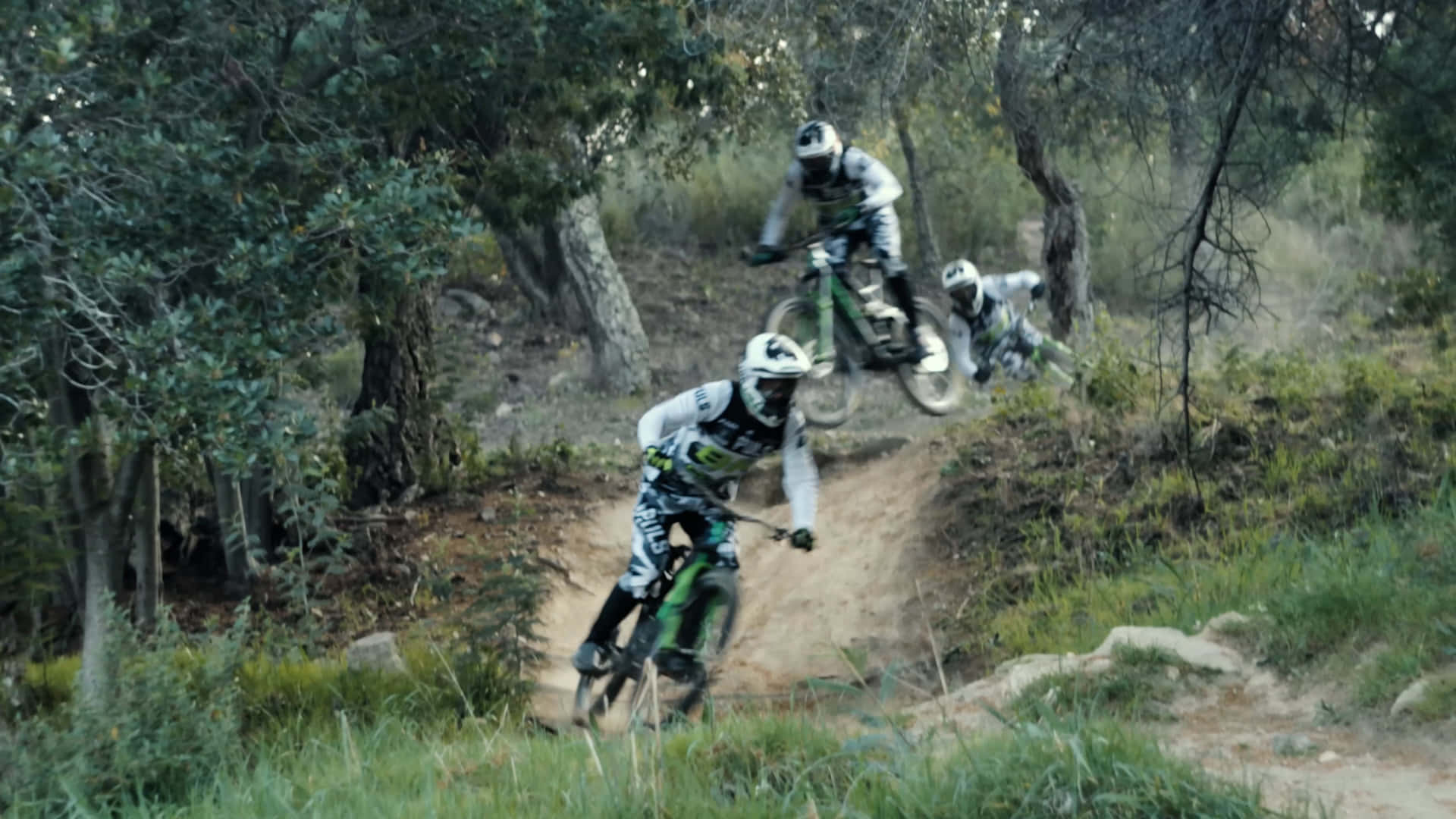bmx 桌布 壁紙 — 三名登山車騎士在塵土飛揚的森林小徑上疾馳而下，在穿越陡峭的樹林下坡時揚起泥土。