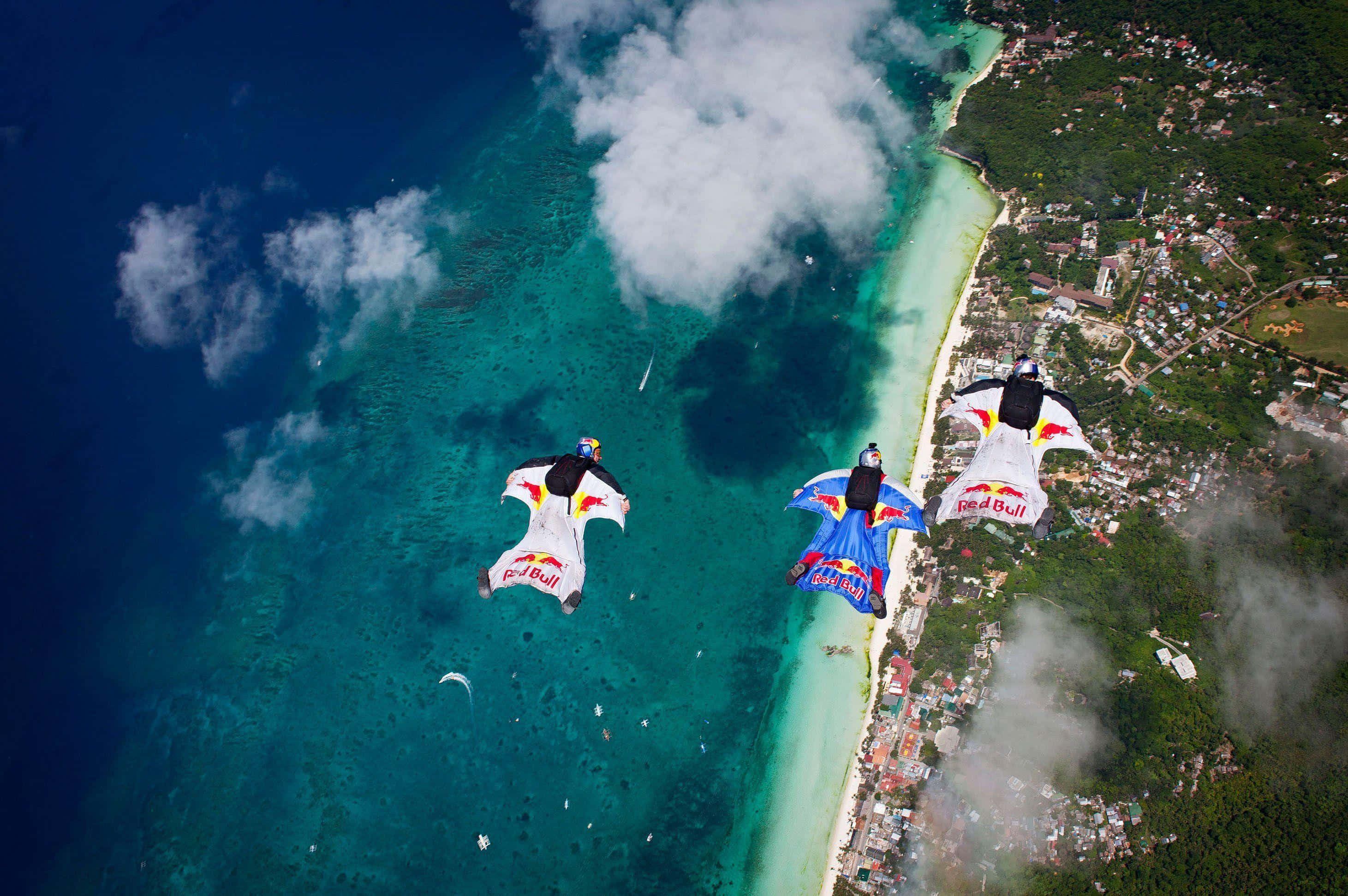 Skydiving 桌布 壁紙 — 三名穿著Red Bull翼裝的跳傘者在碧綠海水和茂密熱帶海岸線上空翱翔。