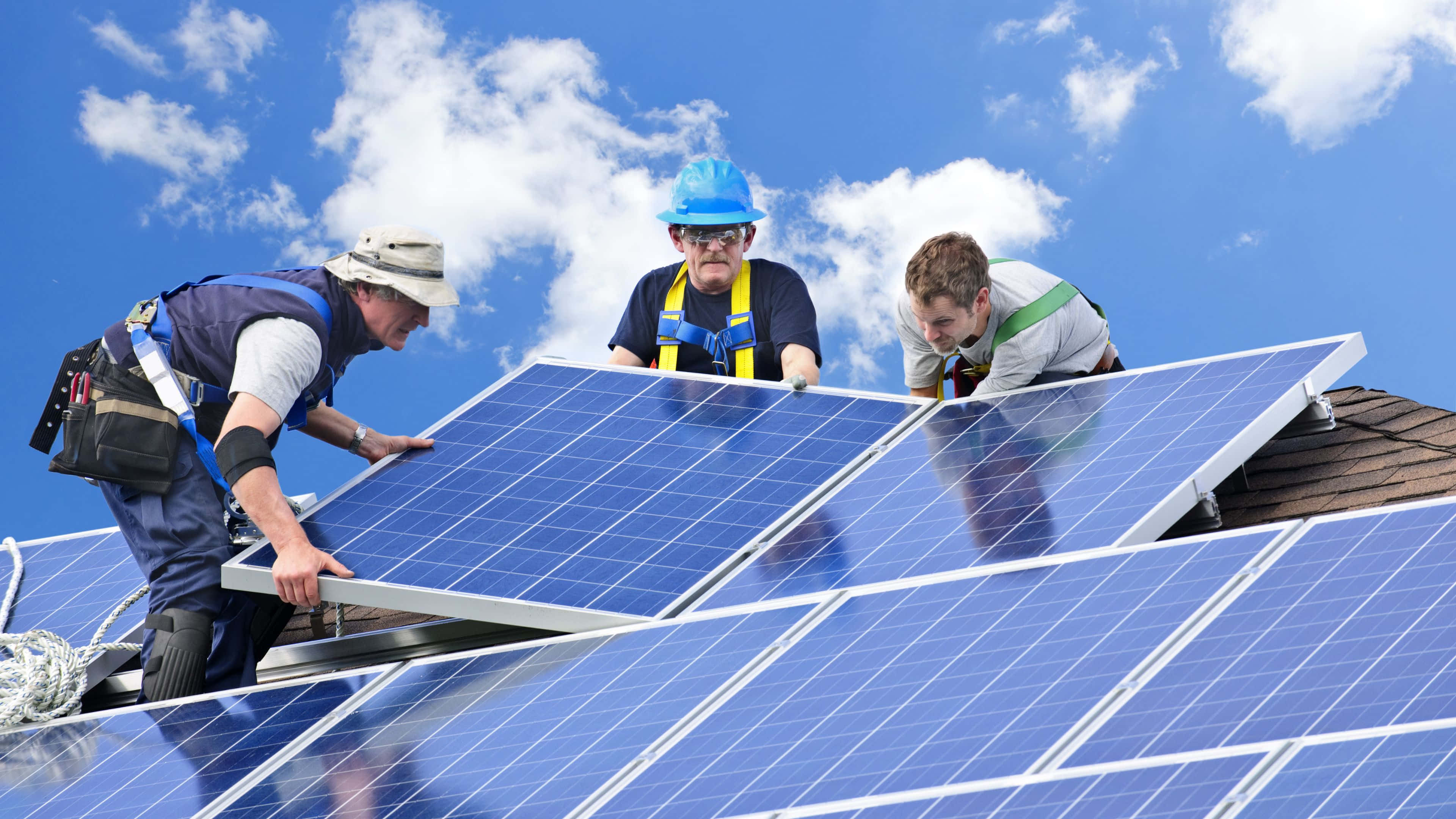 Solar Panel 桌布 壁紙 — 三名穿著安全裝備的工人在晴朗藍天白雲下，在屋頂上安裝藍色太陽能板。