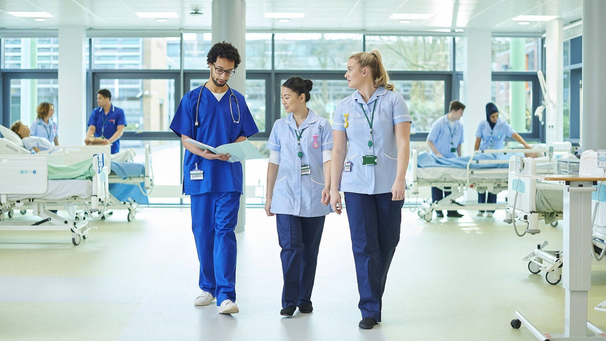 Hospital 桌布 壁紙 — 三名穿著手術衣的醫護人員走過一個明亮現代、有病人躺在床上的醫院病房。