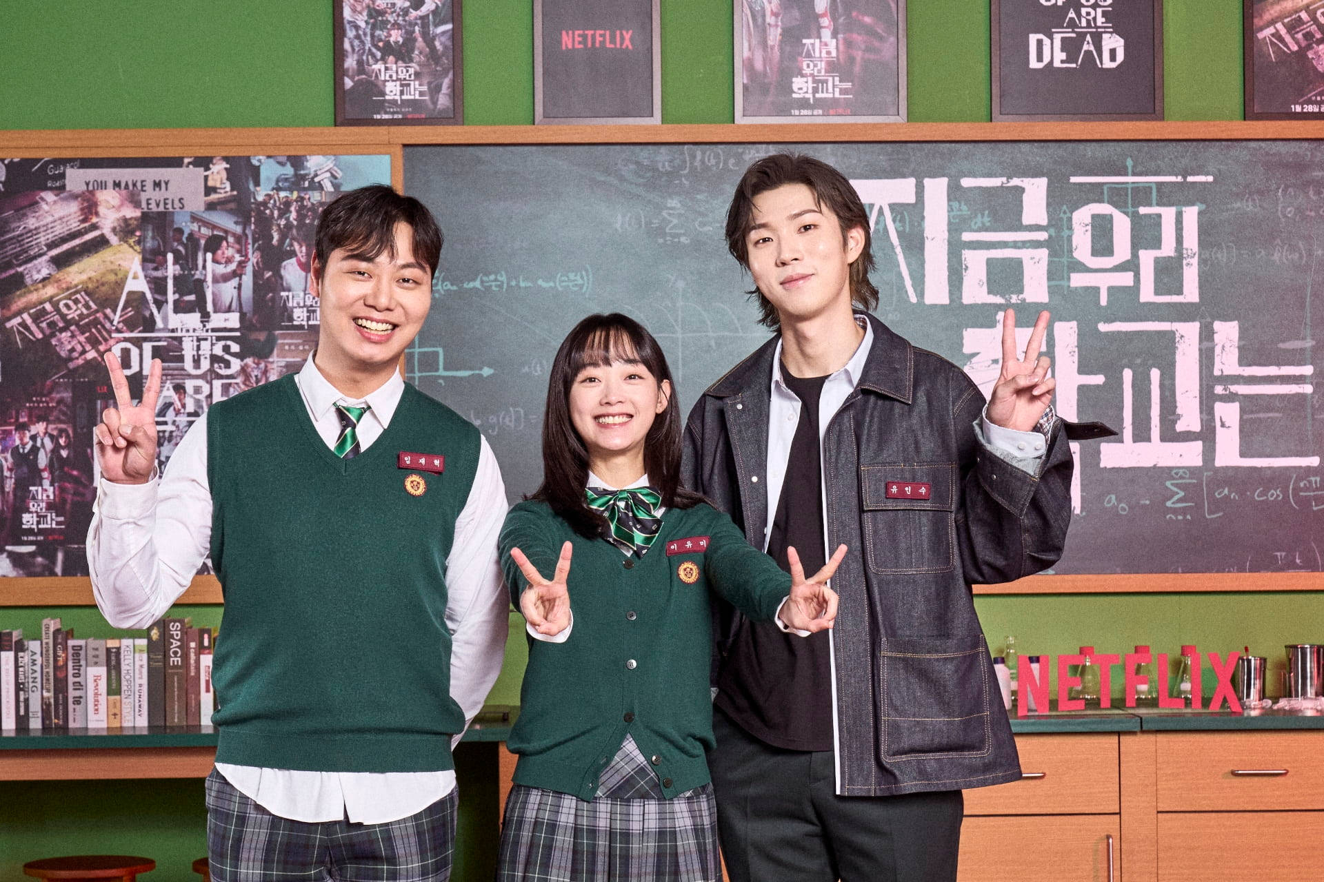 Netflix 桌布 壁紙 — 三名穿著綠色校服的學生面帶微笑，在裝飾著Netflix海報的教室裡比出和平手勢。