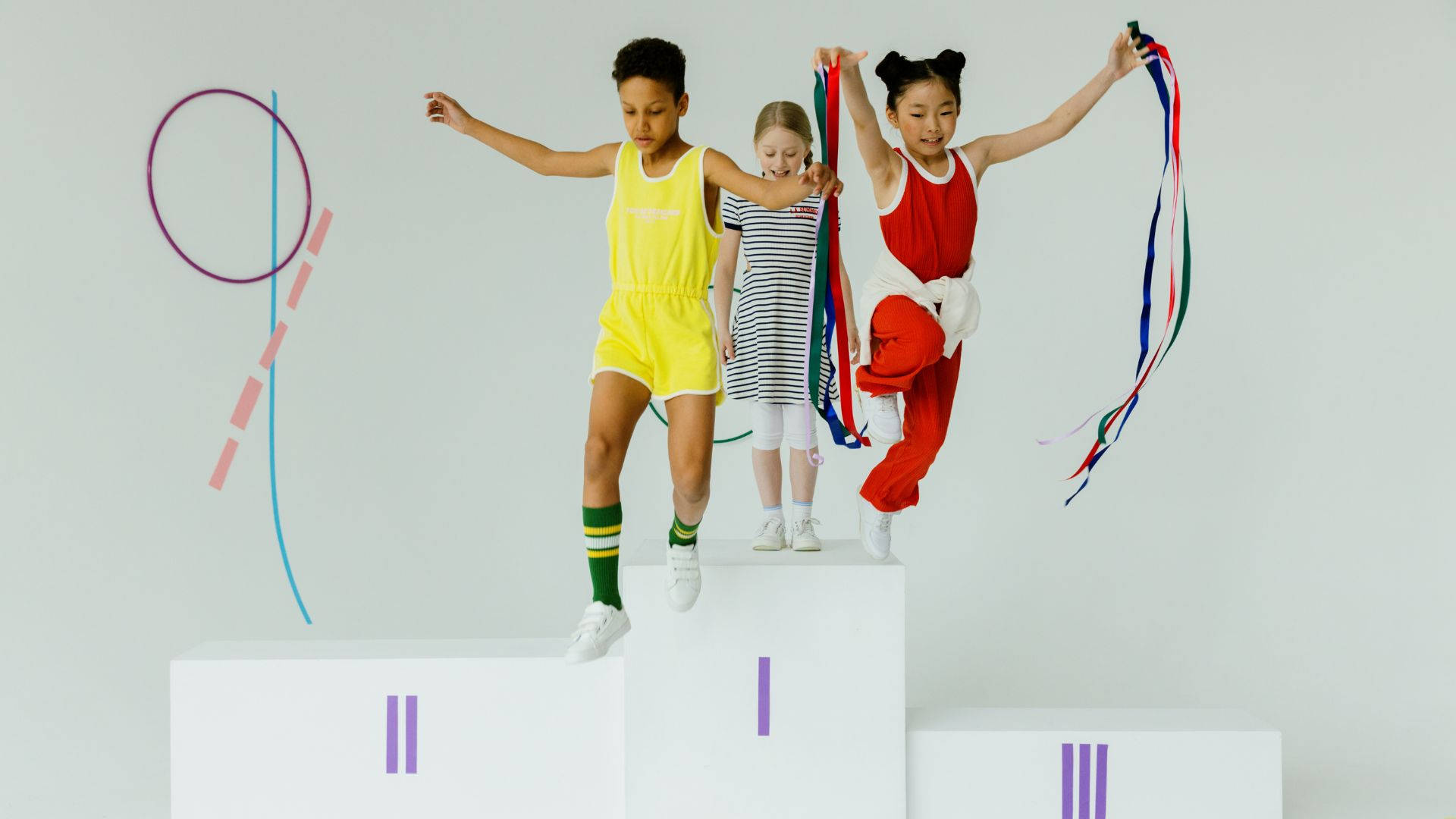 Olympics 桌布 壁紙 — 三名穿著運動服的孩子在簡約的白色頒獎台上，以抽象彩色圖案為背景的素牆前歡樂慶祝。