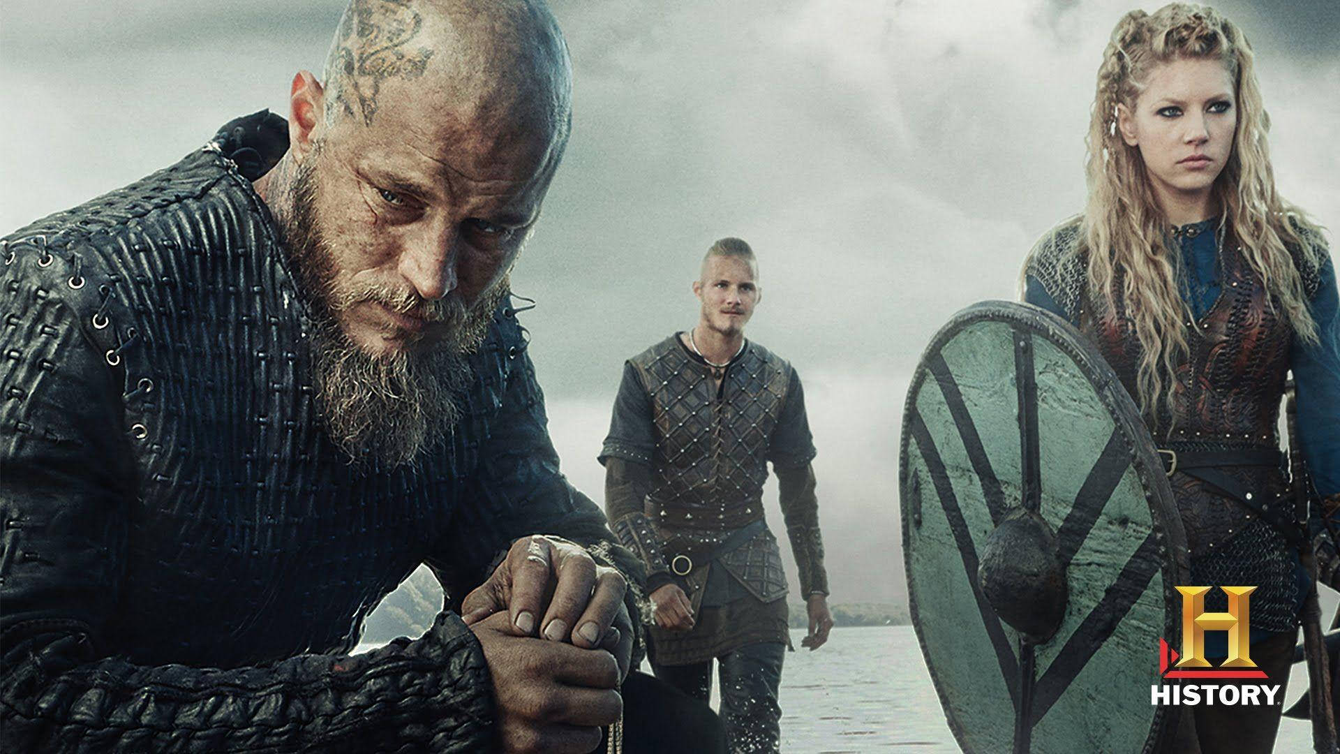 Vikings 桌布 壁紙 — 三名維京戰士在暴風雨的天空下莊嚴地站在水邊。一人握著盾牌，另一人專注地凝視前方。