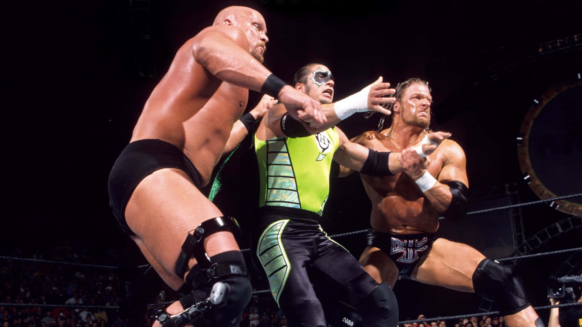triple h 桌布 壁紙 — 三名肌肉發達的摔角手在明亮的場館燈光下，在擂台上激烈纏鬥，背景是模糊的人群。