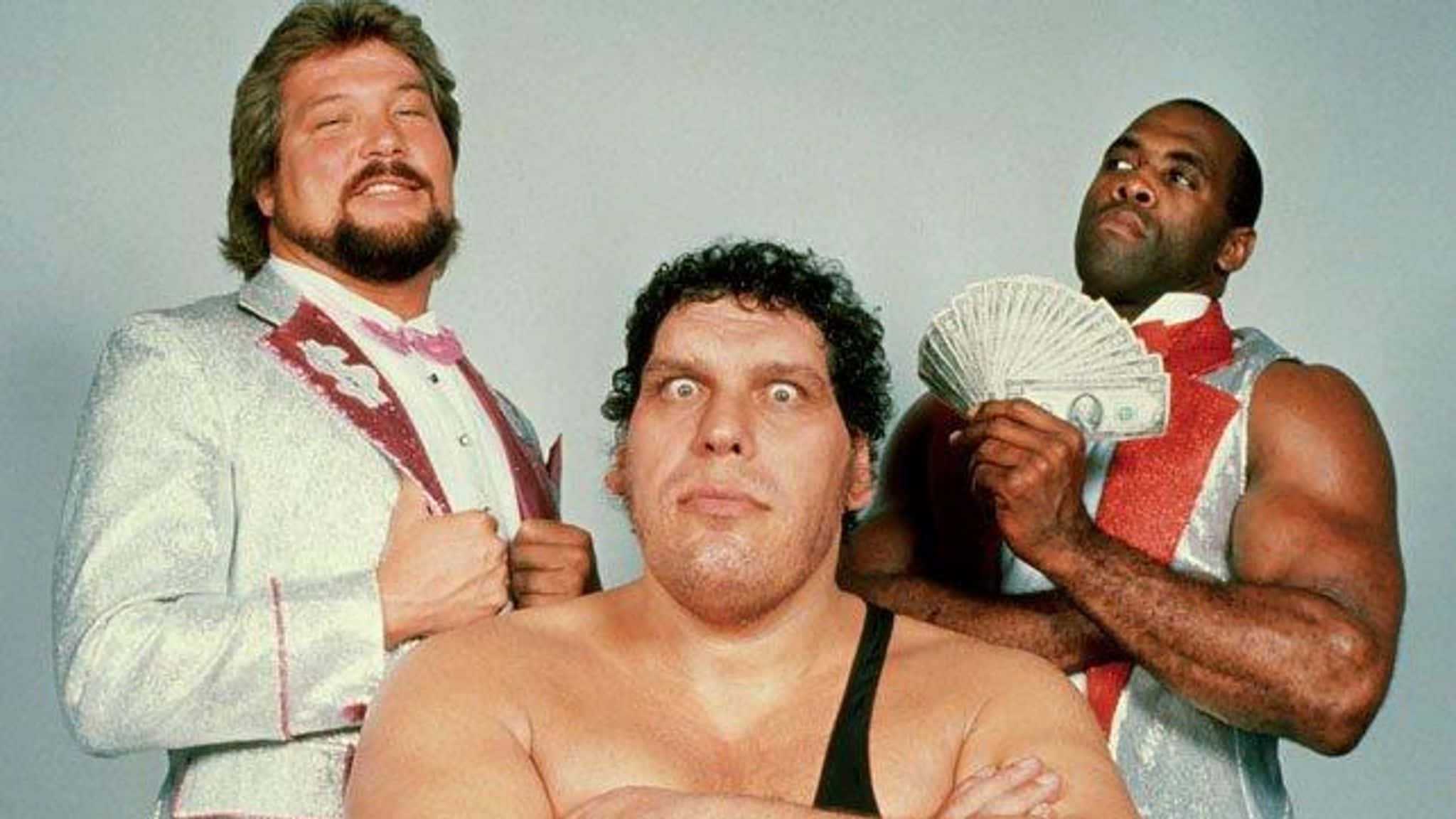 Andre The Giant 桌布 壁紙 — 三名肌肉發達的摔角手在純灰色背景下戲劇性地擺姿勢，一人手持扇狀現金，另一人穿著閃亮西裝配蝴蝶結。