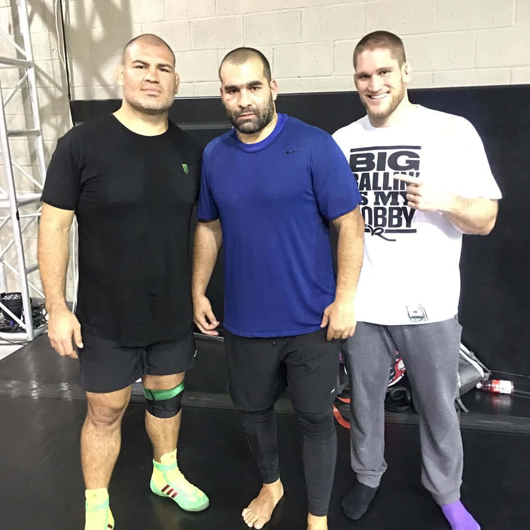 MMA 桌布 壁紙 — 三名肌肉發達的男子穿著運動服，在黑色墊子上自信地擺姿勢，背景是煤渣磚牆和舞台設備。