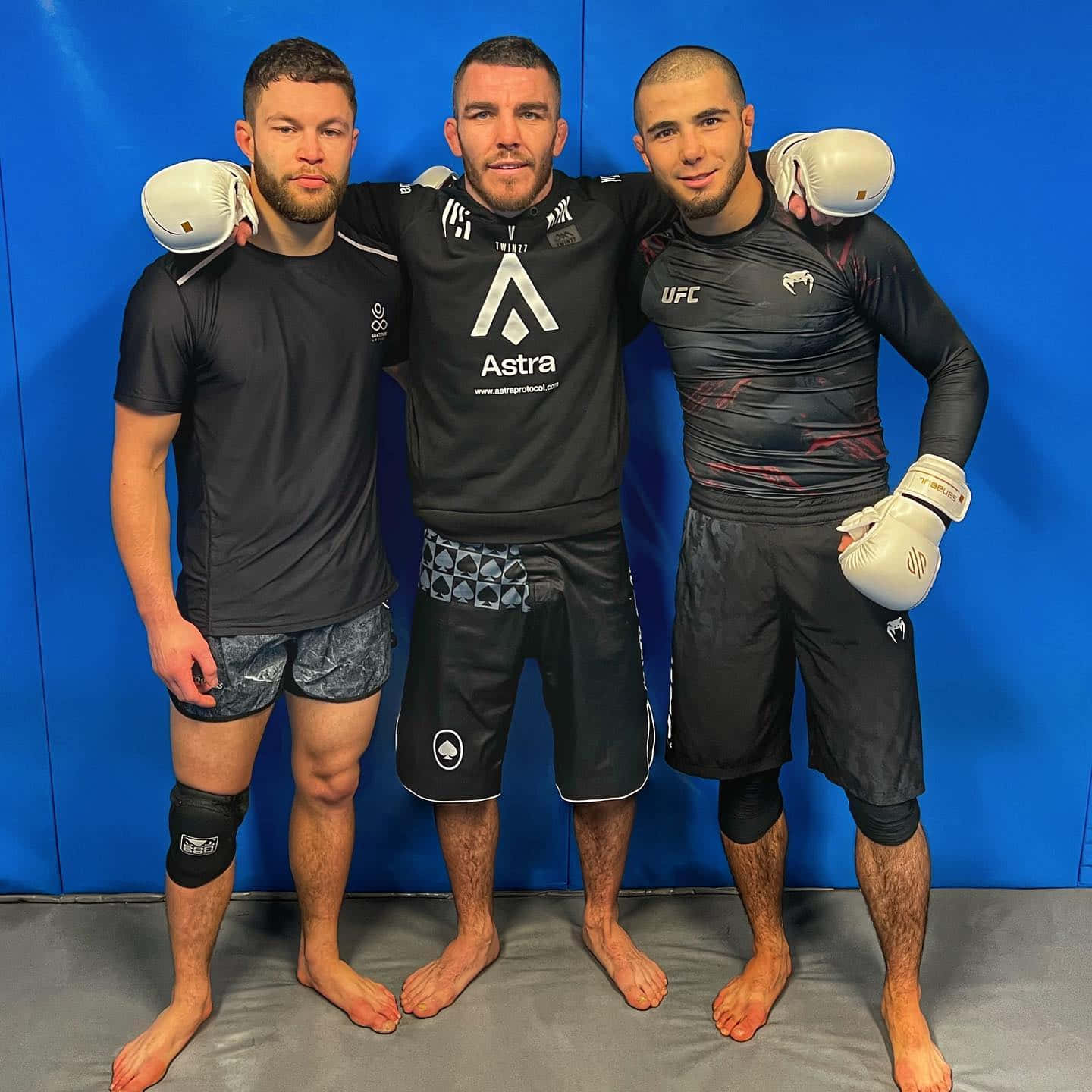UFC 桌布 壁紙 — 三名赤腳男性拳手身穿訓練裝備，自信地互相搭肩，靠著藍色軟墊牆壁擺姿勢。