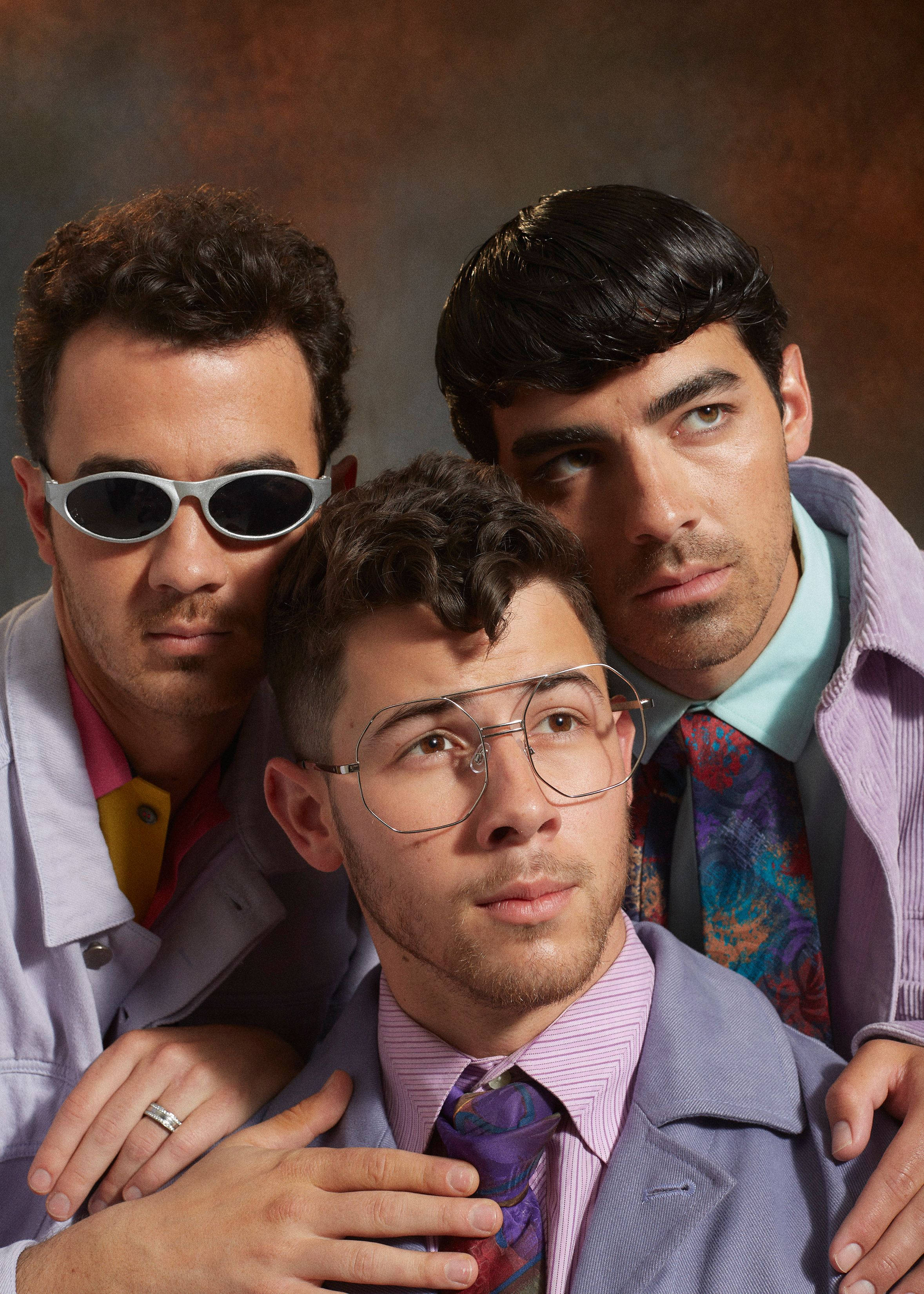 Jonas Brothers 桌布 壁紙 — 三名身穿1970年代風格服裝、戴著太陽眼鏡和圖案領帶的男子，在溫暖的棕色背景前緊密地擺姿勢。
