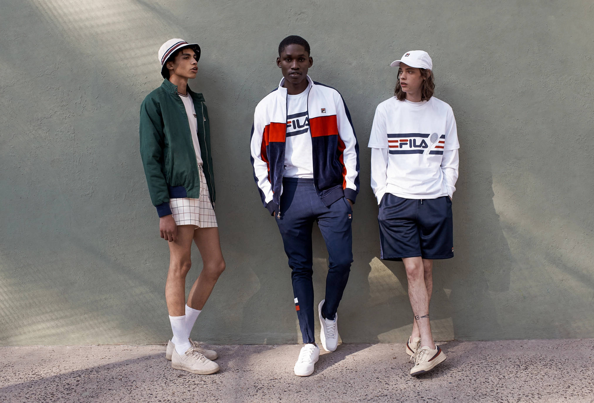 Fila 桌布 壁紙 — 三名身穿Fila運動服的年輕男子在明亮的日光下隨意靠在一面有紋理的灰色牆上。