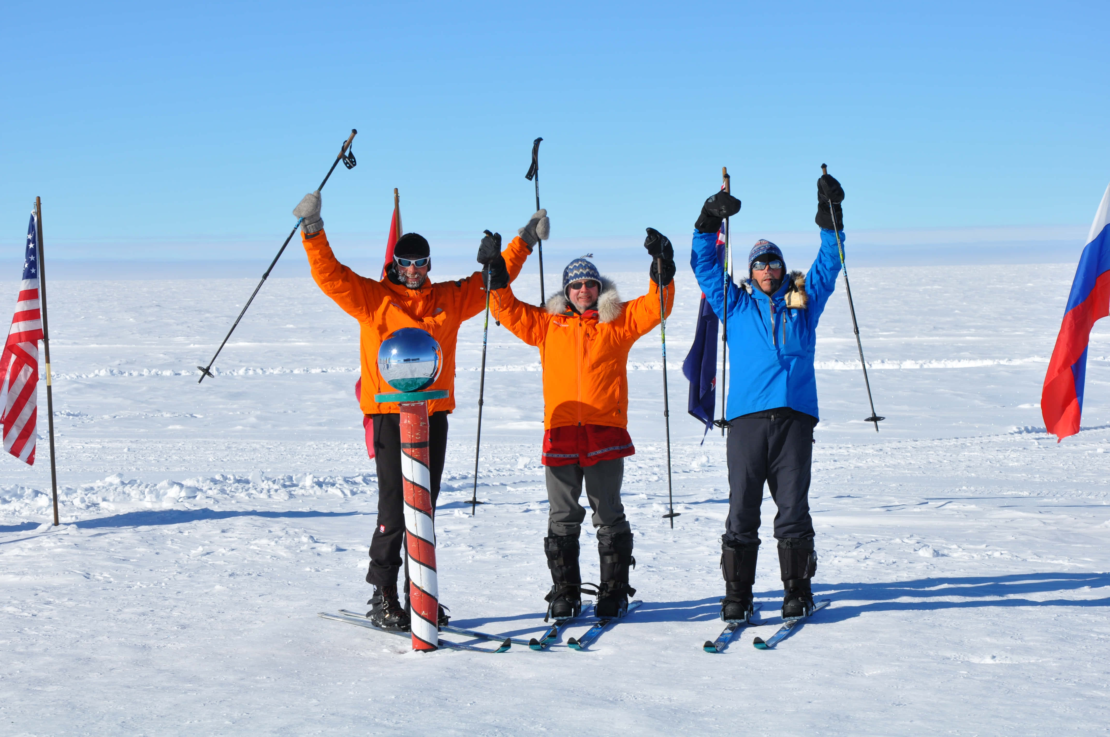 South Pole 桌布 壁紙 — 三名身穿亮色外套的滑雪者在晴朗藍天下廣闊的雪原上勝利慶祝，身旁有旗幟。